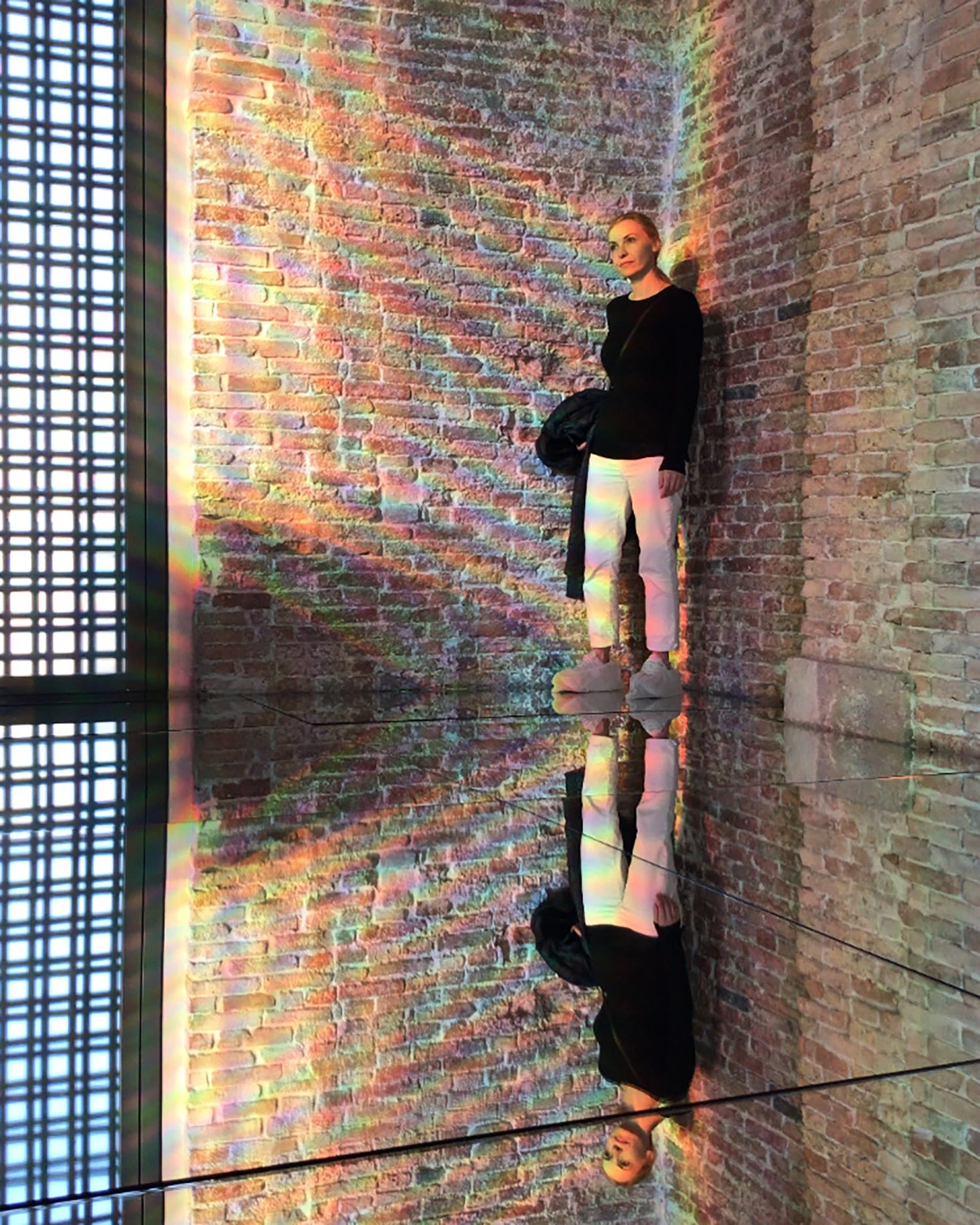 Visual artist Aggeliki Dimitriadou standing in an art installation with rainbow light reflections on a brick wall and a mirrored floor.