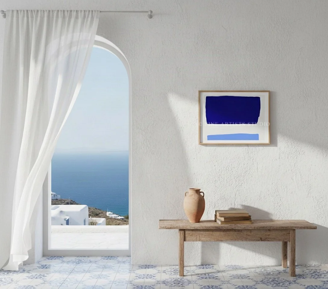 Framed modern abstract geometric print in cobalt and sky blue hanging on a neutral wall above a wooden table with organic ceramic stools and decorative vases.