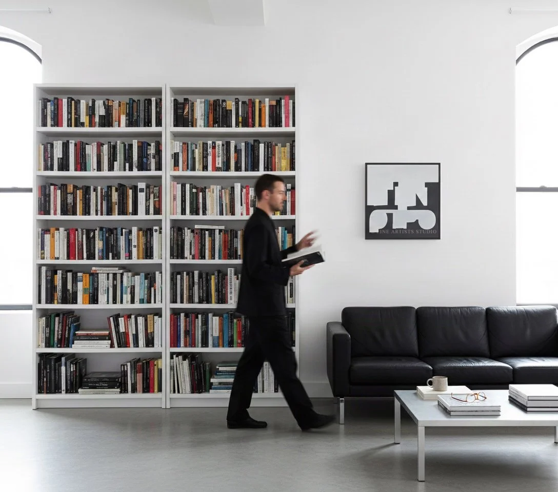 Contemporary room interior with the square painting "Signal III" hanging next to a tall white double bookcase filled with books above a black leather sofa.