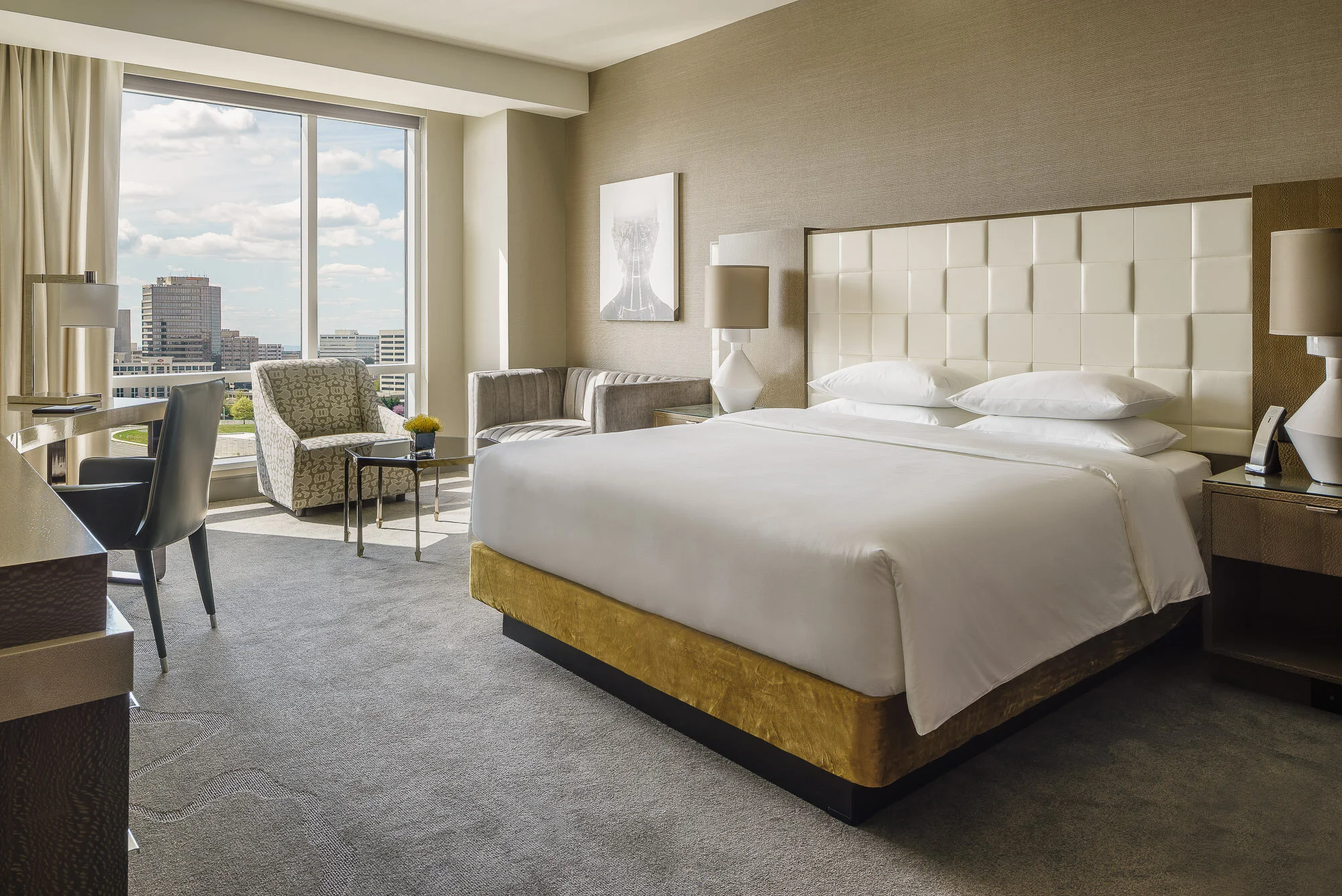 Modern hotel room with a king-size bed, large window with city view, two chairs, small table, and neutral decor.