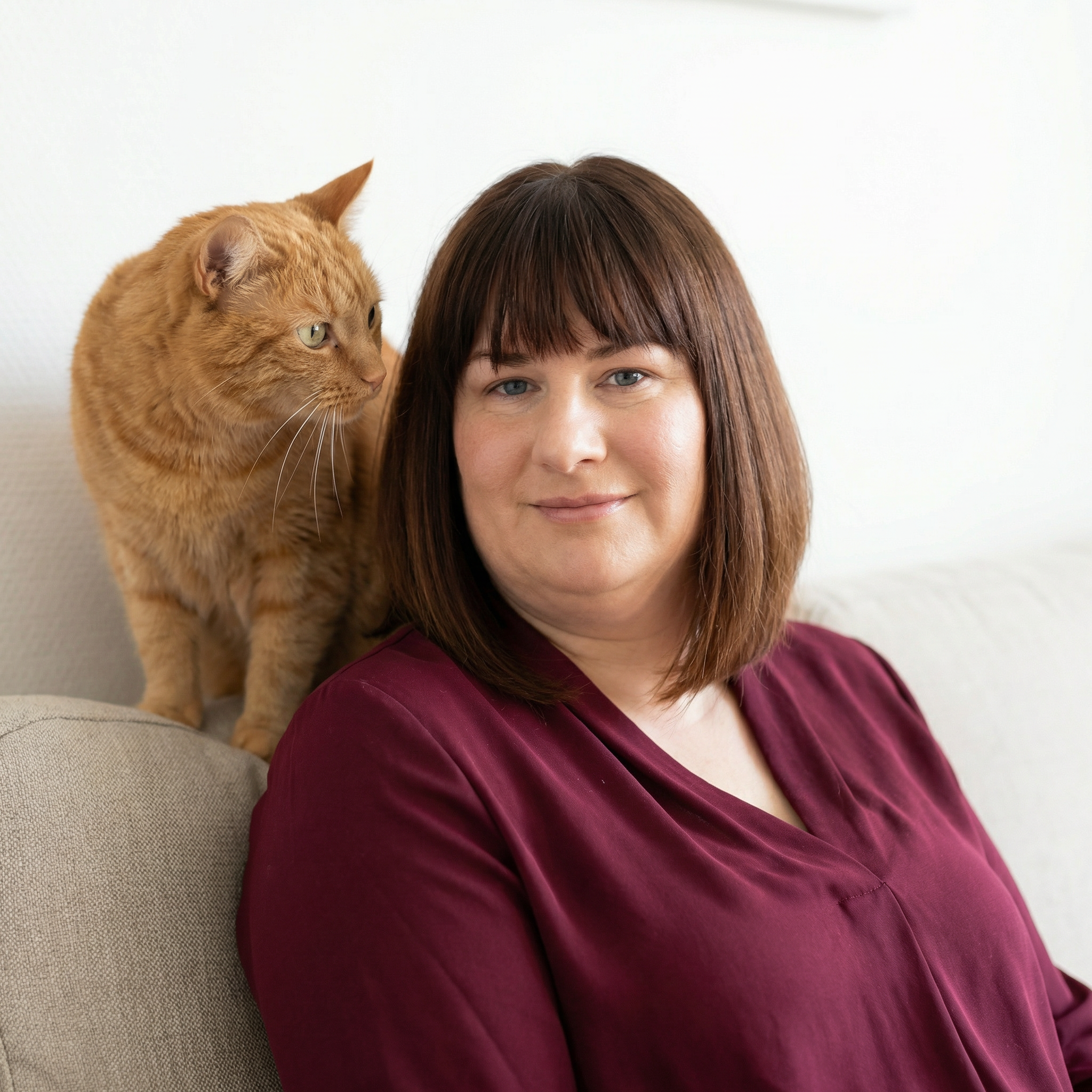 Une femme aux cheveux bruns avec un chat orange sur l'épaule, assise sur un canapé beige contre un mur blanc.