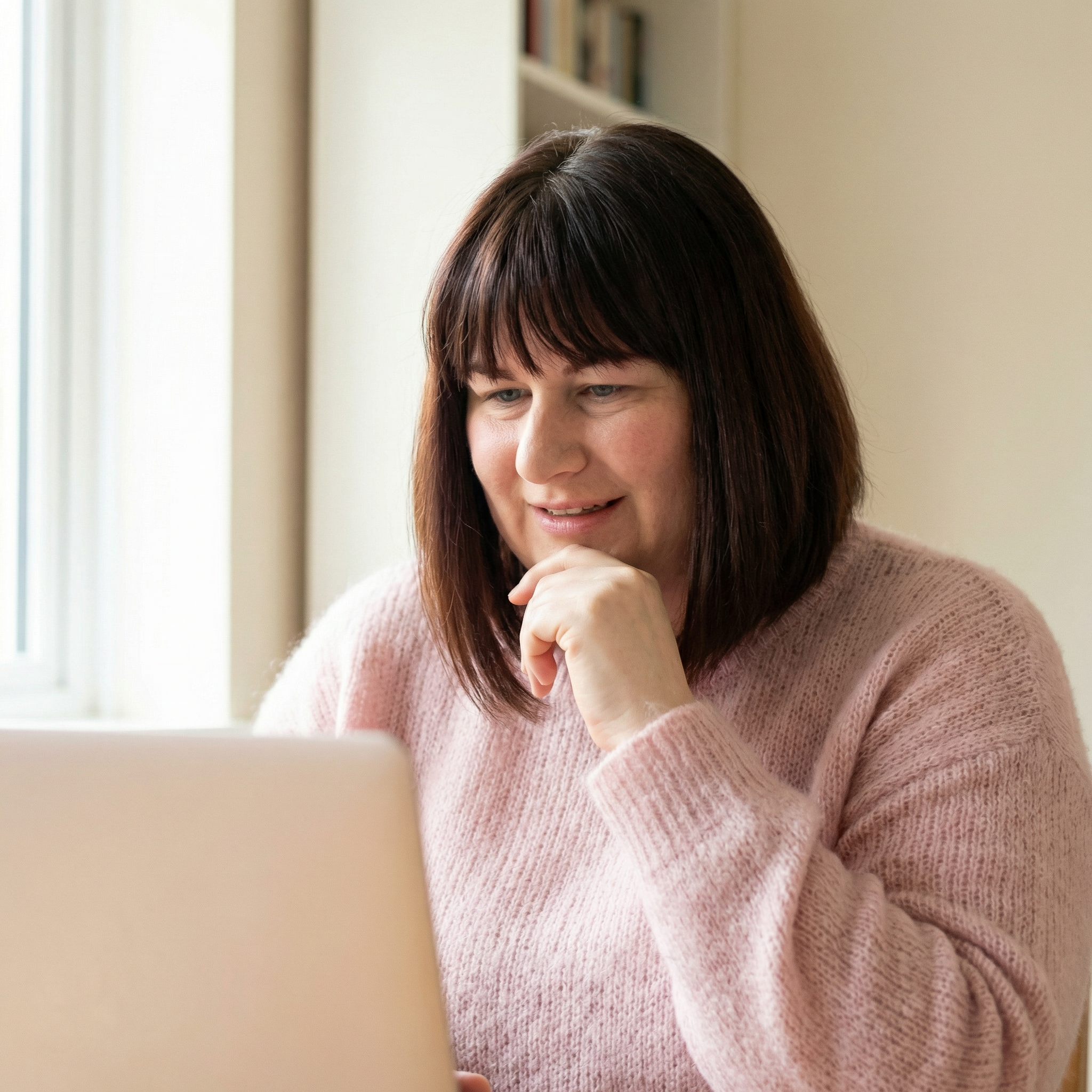 Femme avec cheveux bruns et coupe en dégradé portant un pull rose clair, utilisant un ordinateur portable dans une pièce lumineuse.