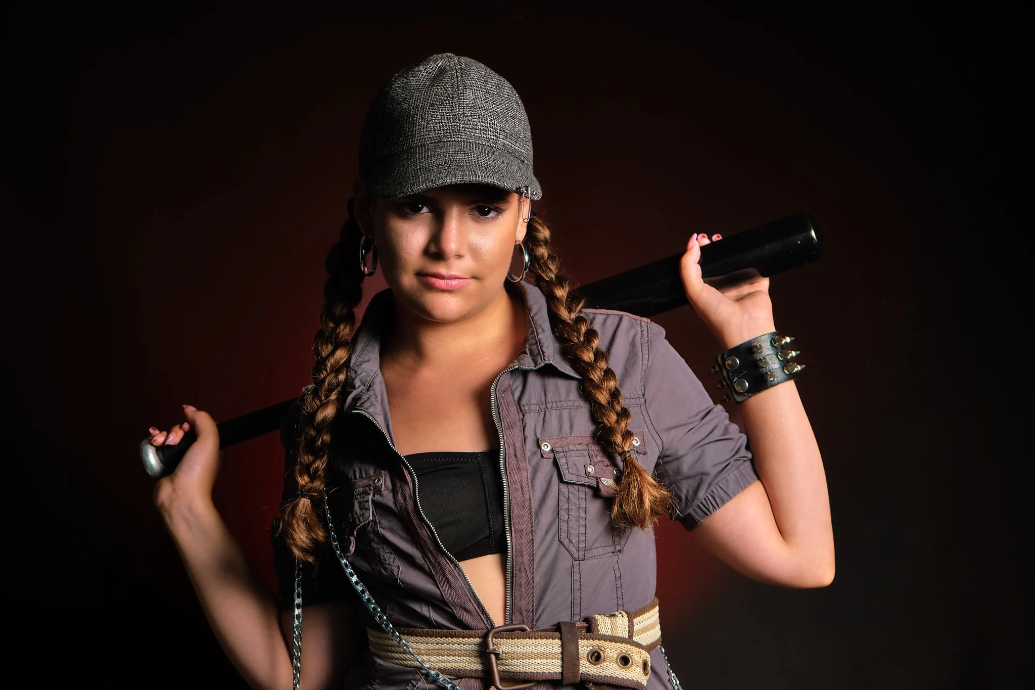 Joven mujer con gorra, ropa de estilo urbano, con una especie de cinturón en la cintura y una pulsera con púas, sosteniendo un bate de béisbol sobre sus hombros, de fondo oscuro.