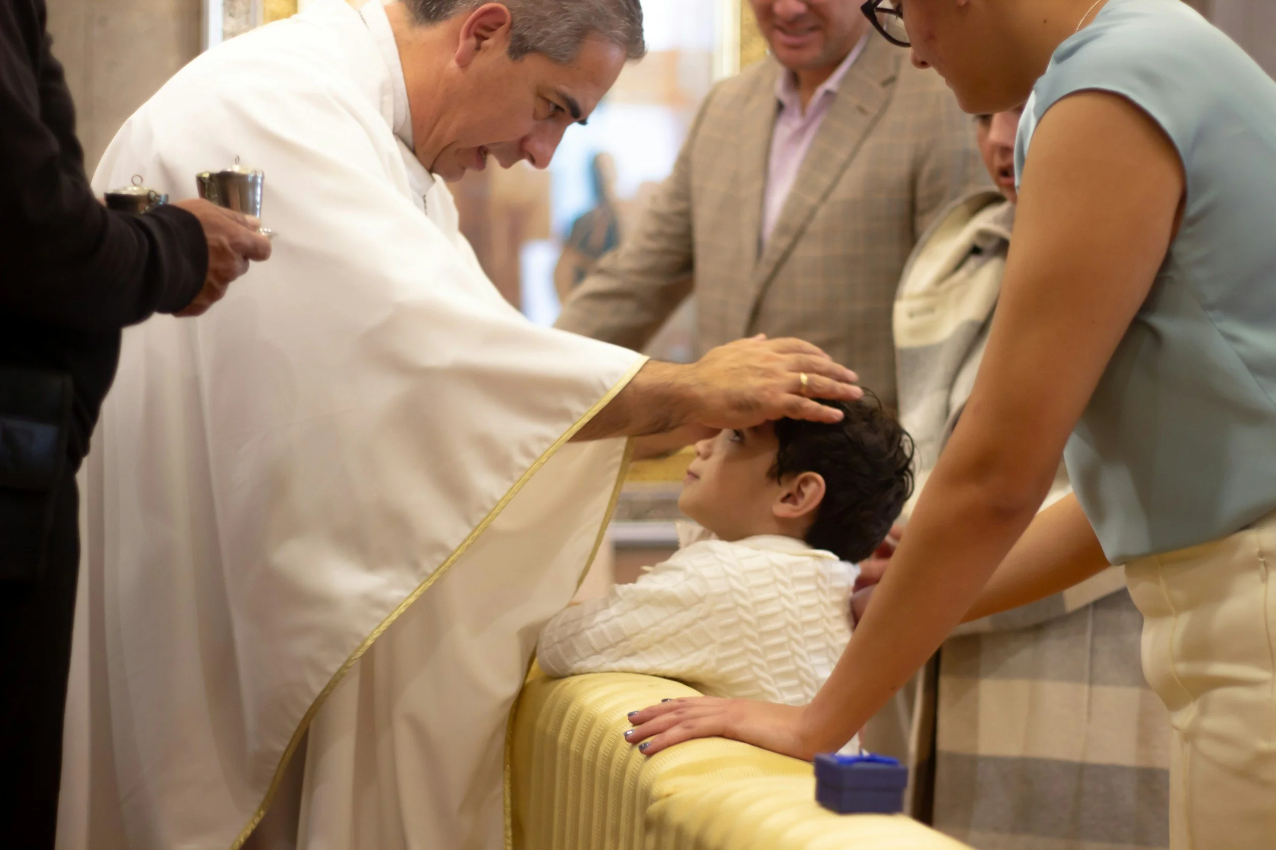 Un sacerdote bendiciendo a un niño sentado en una banca, mientras otras personas observan y participan en la ceremonia.
