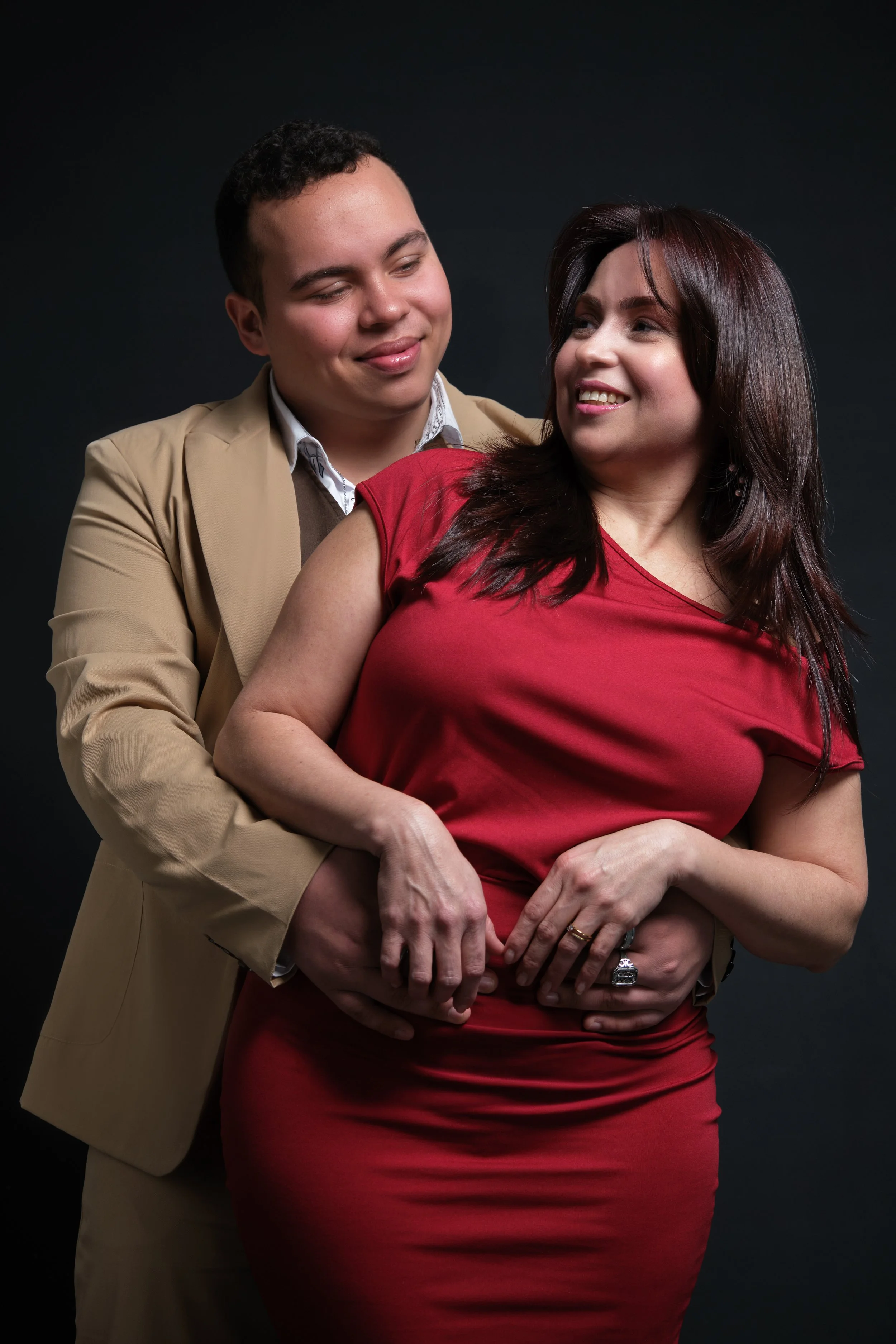 Una pareja posando juntos, el hombre abraza a la mujer por la cintura y ambos sonríen, con fondo negro.