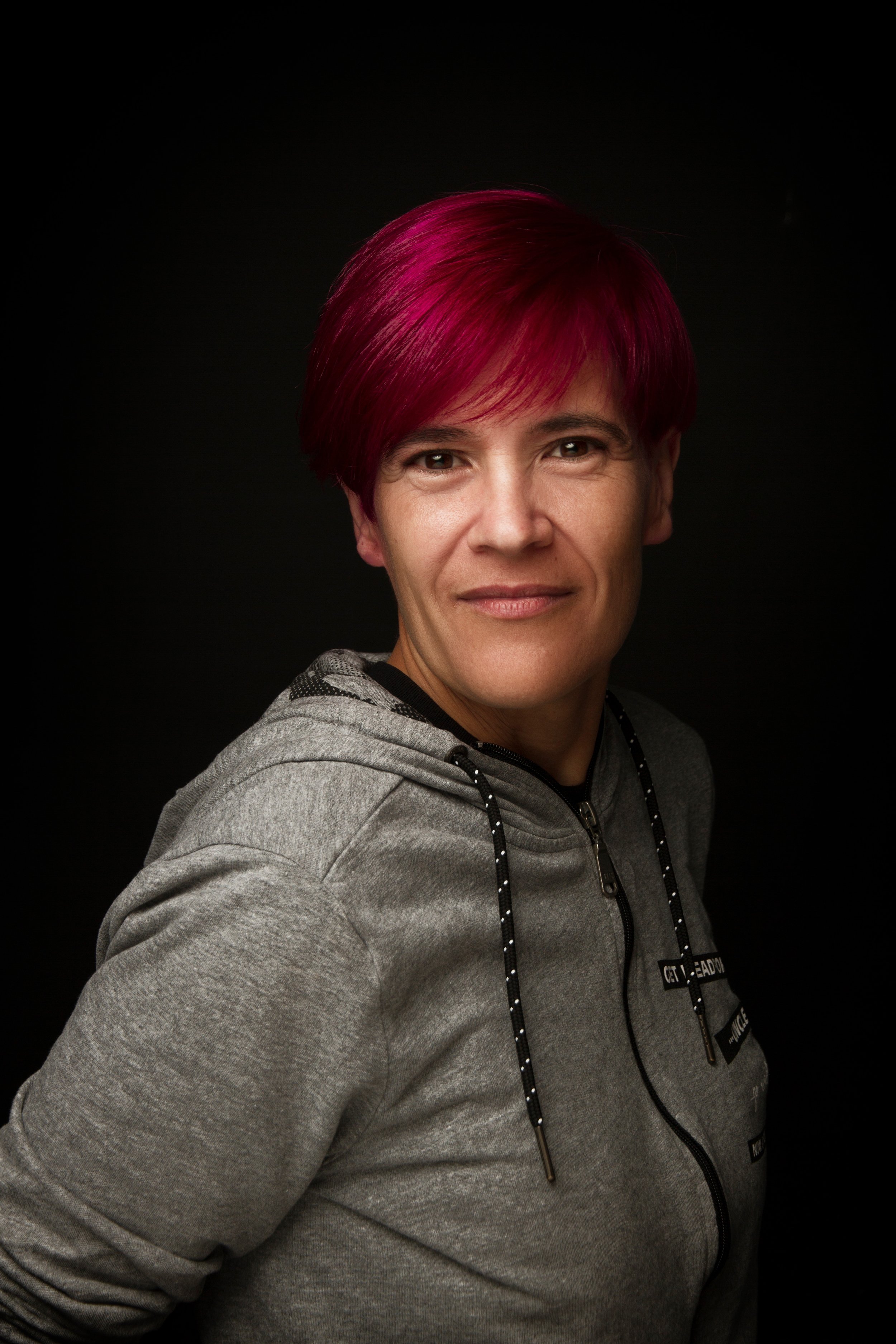 Retrato de una mujer con cabello corto, teñido de color rosa, viste sudadera gris y tiene fondo negro.