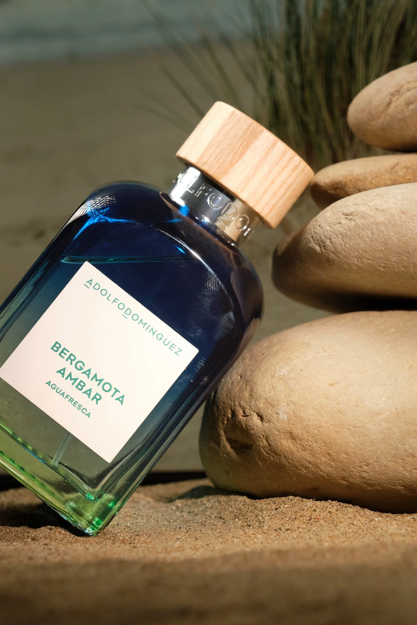 Botella de perfume con etiqueta que dice 'Bergamota Ambar' y 'Aguafresca', con tapón de madera, descansando sobre arena junto a piedras}