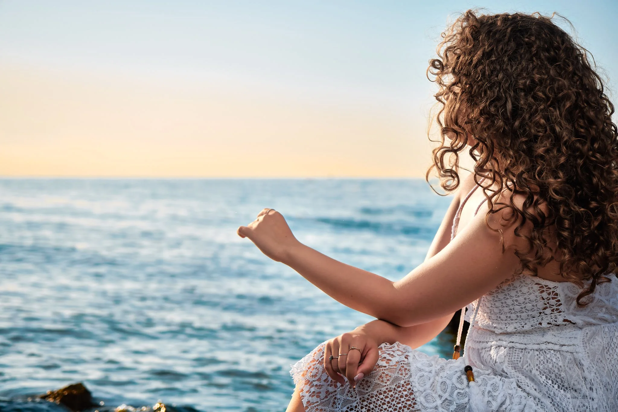 Una mujer con cabello rizado de color castaño claro, usando un vestido de encaje blanco, sentada cerca del mar durante la puesta del sol.