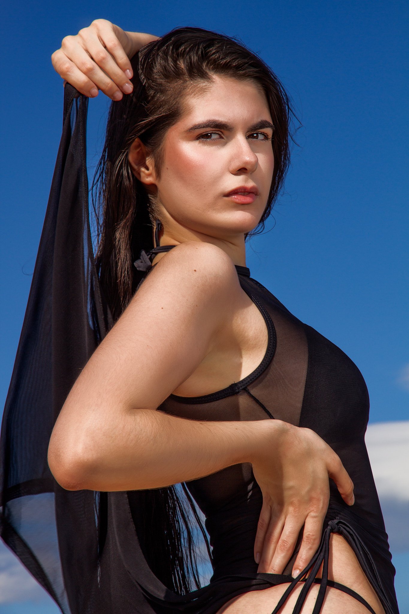 Una mujer con cabello oscuro y piel clara, vestida con ropa negra de encaje, posando al aire libre contra un cielo azul con algunas nubes.