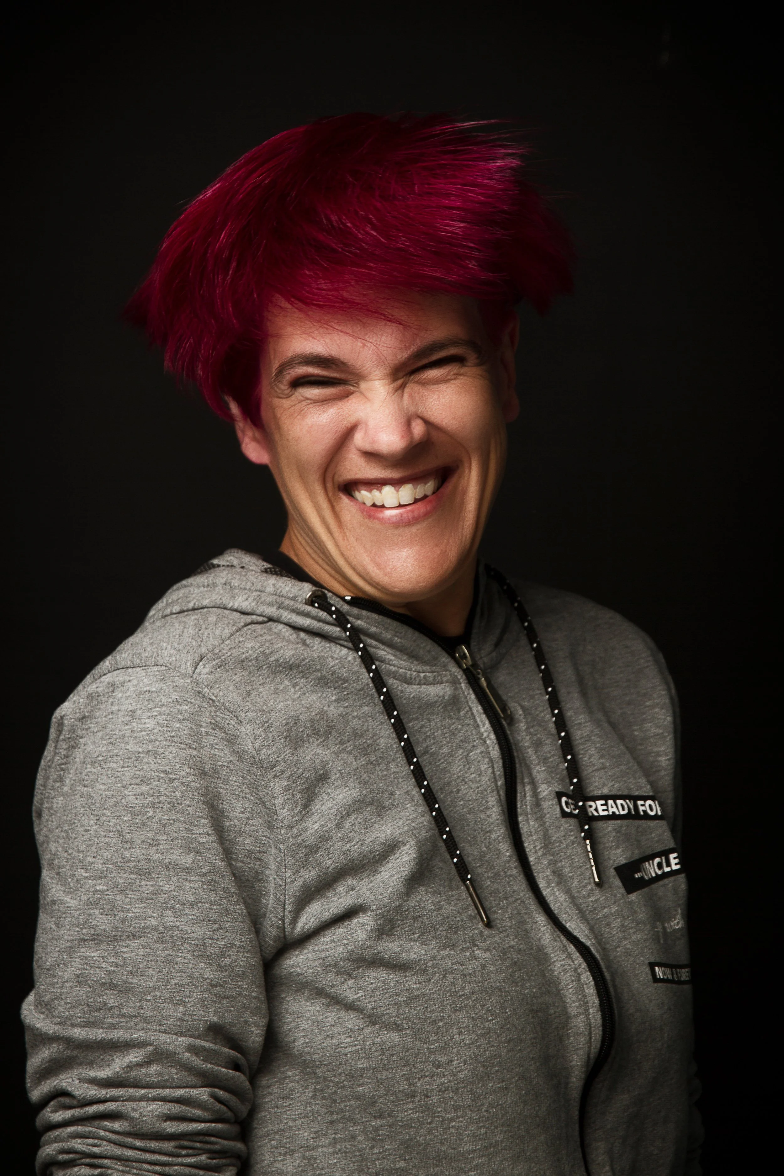 Una mujer sonriendo con cabello corto y teñido de rosa, lleva una sudadera gris con cremallera y fondo negro.