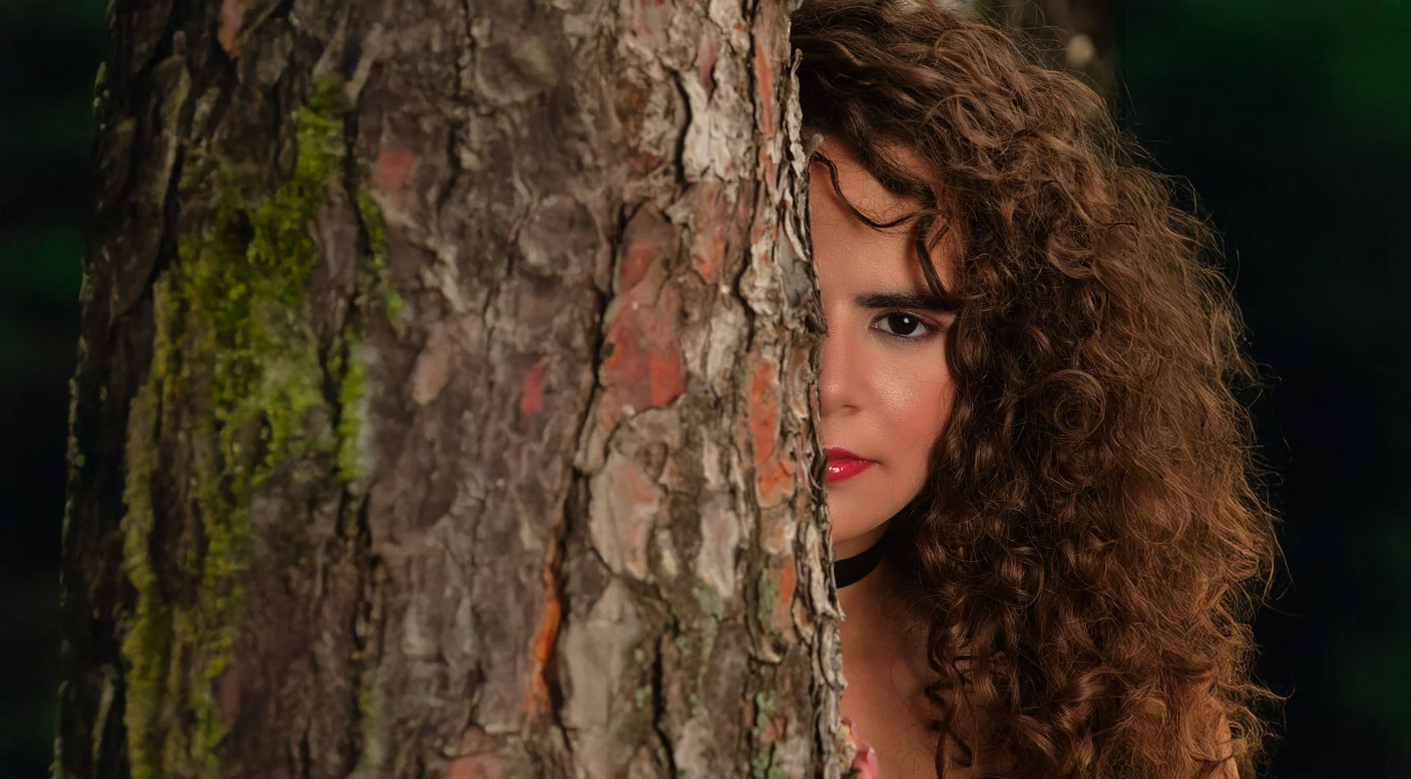Mujer con cabello rizado mirando desde detrás de un árbol en un entorno natural.