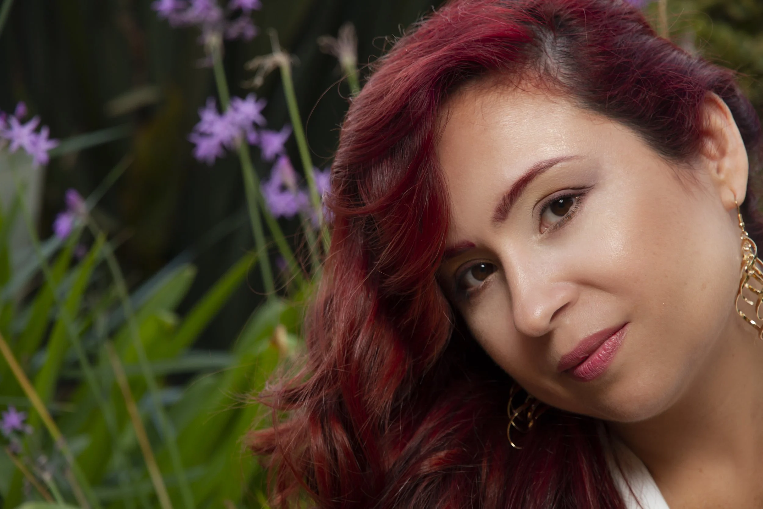Retrato de una mujer con cabello rizado y rojo, en un entorno natural con flores moradas y verdes.