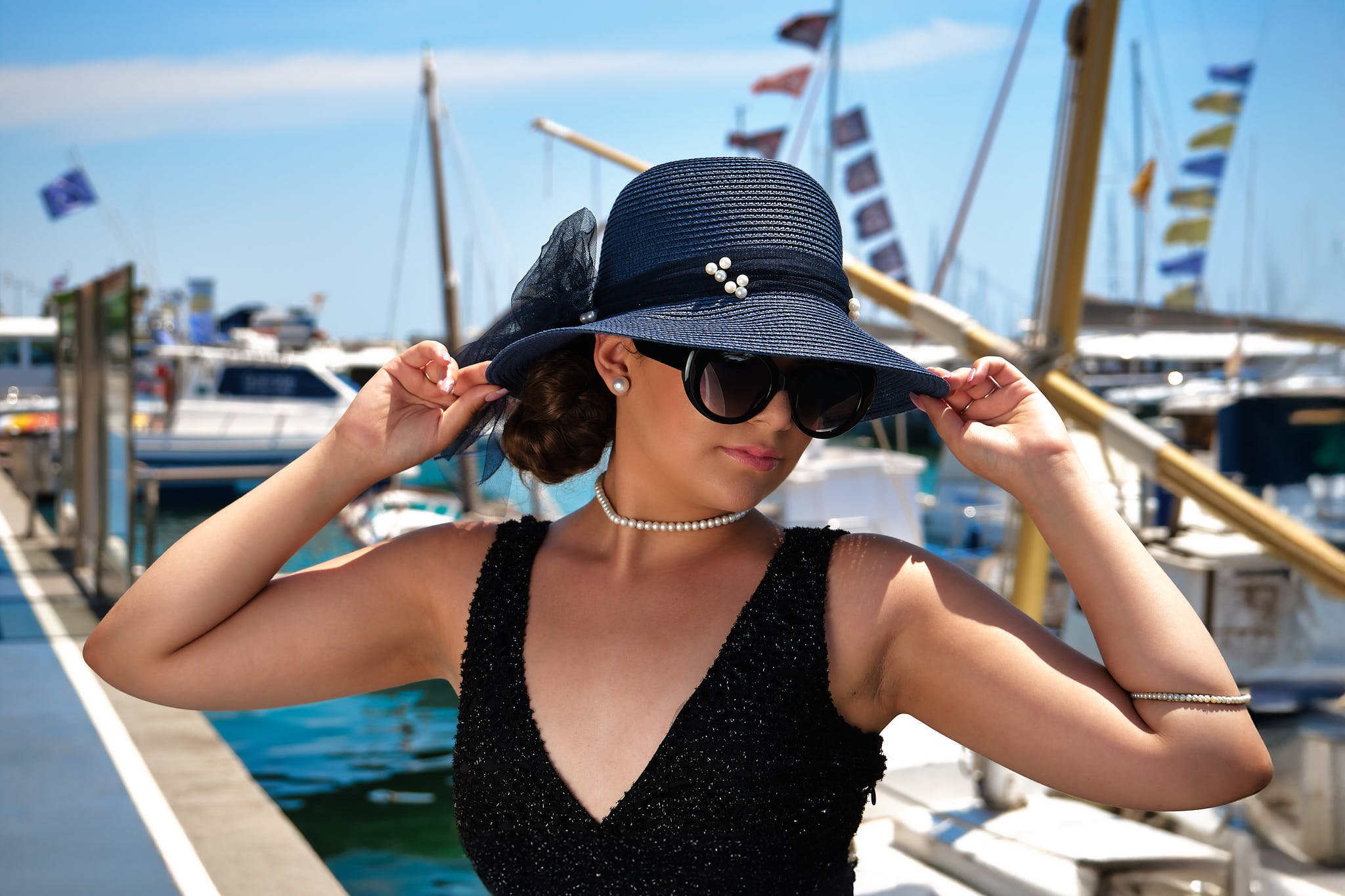 Mujer con sombrero, gafas de sol y vestido negro en un puerto con barcos.