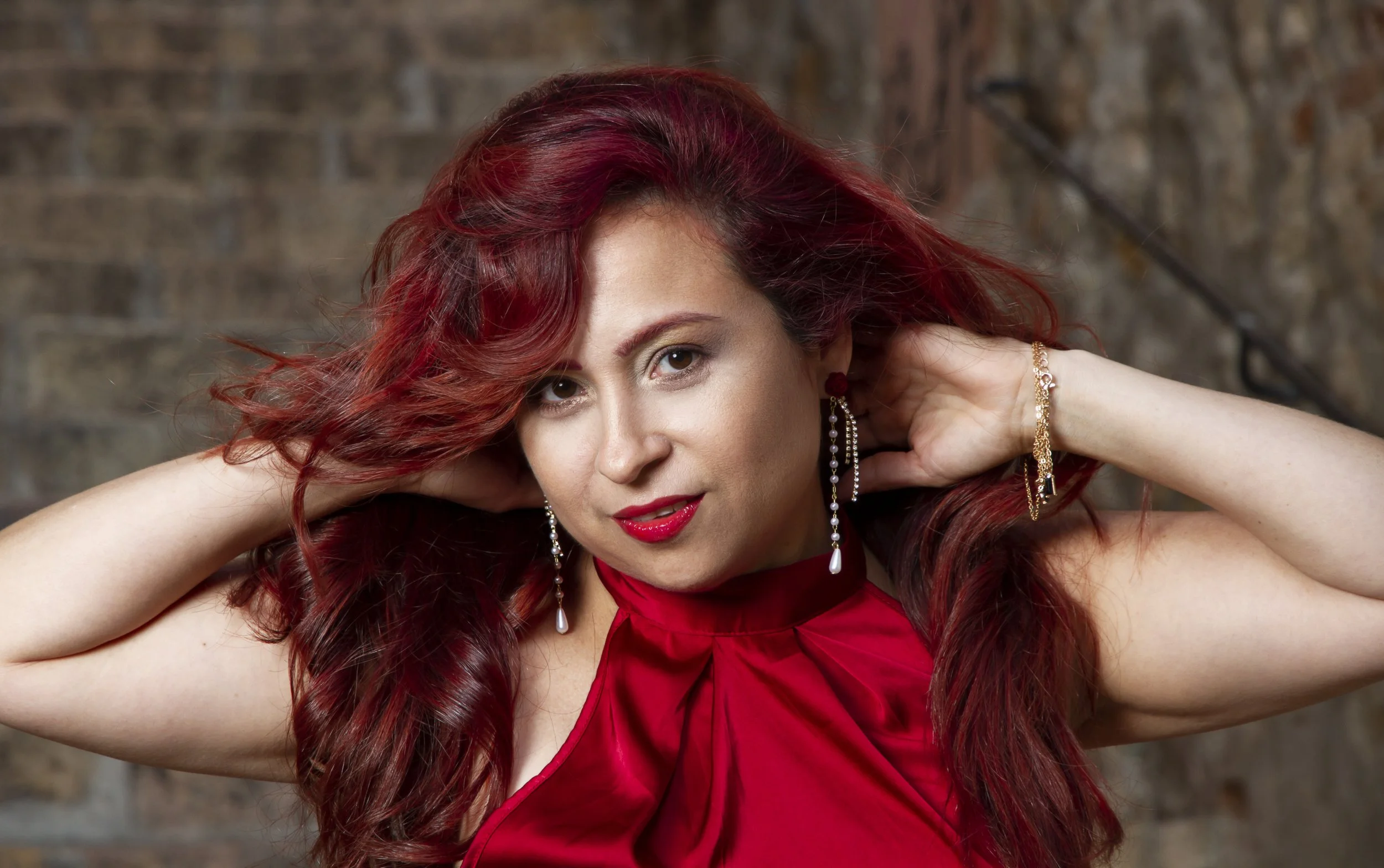 Mujer con cabello rizado y rojo, usando vestido rojo y pendientes largos, frente a una pared de ladrillos.