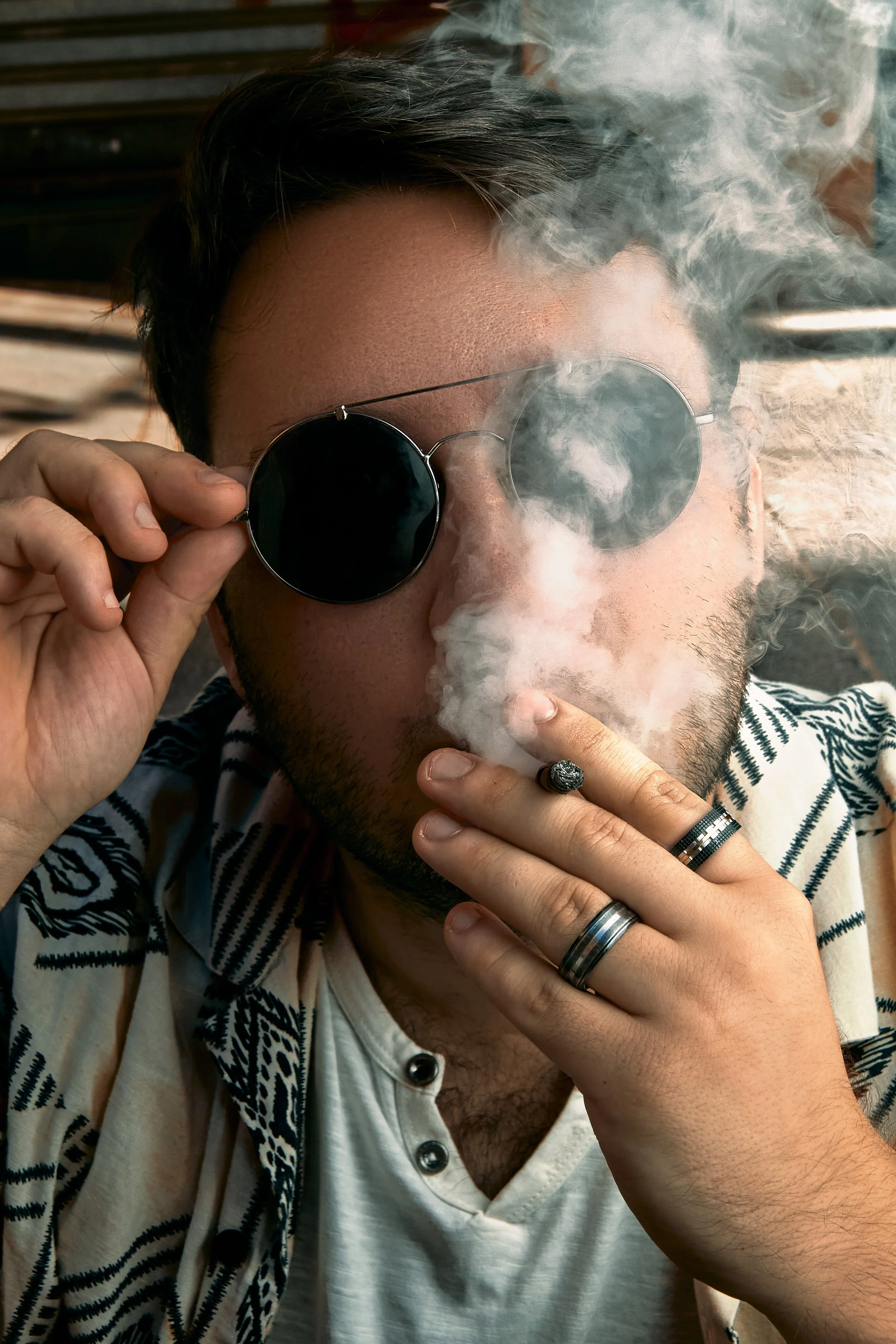 Un hombre con gafas de sol negras, barba ligera, usando una camiseta blanca y una camisa de manga larga con patrones, fuma un cigarro mientras ajusta sus gafas, rodeado de humo.