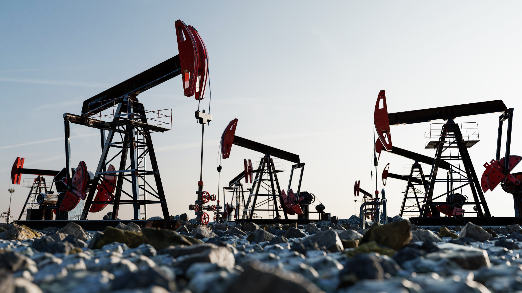 Multiple oil pumpjacks in a field with rocks in the foreground and a clear sky background.
