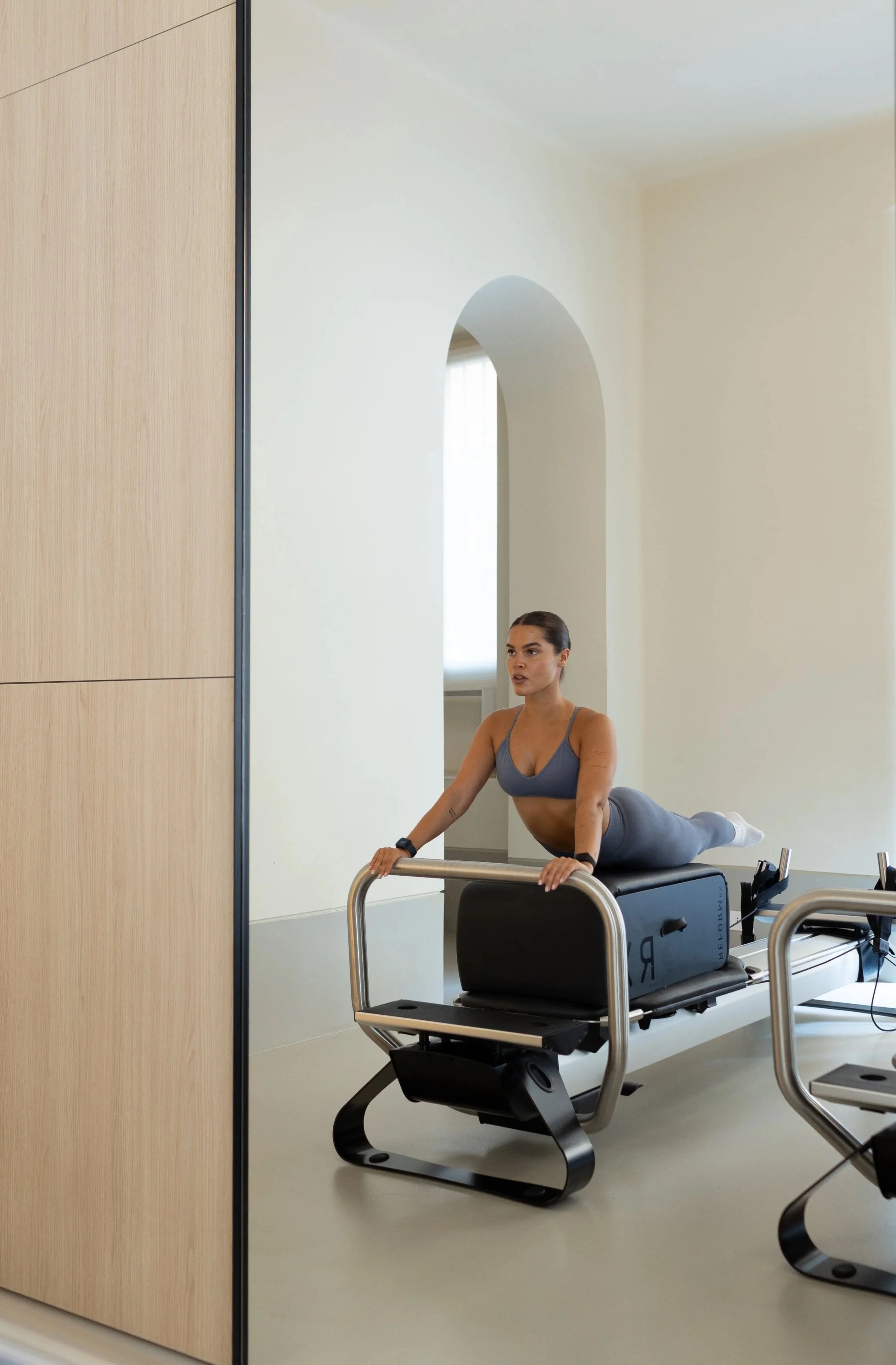 Una donna sta facendo esercizi di yoga su un cuscino imbottito davanti a uno specchio, con una parete in legno sulla sinistra e una parete bianca con una nicchia nell'angolo. Indossa abbigliamento sportivo grigio e sembra concentrata.