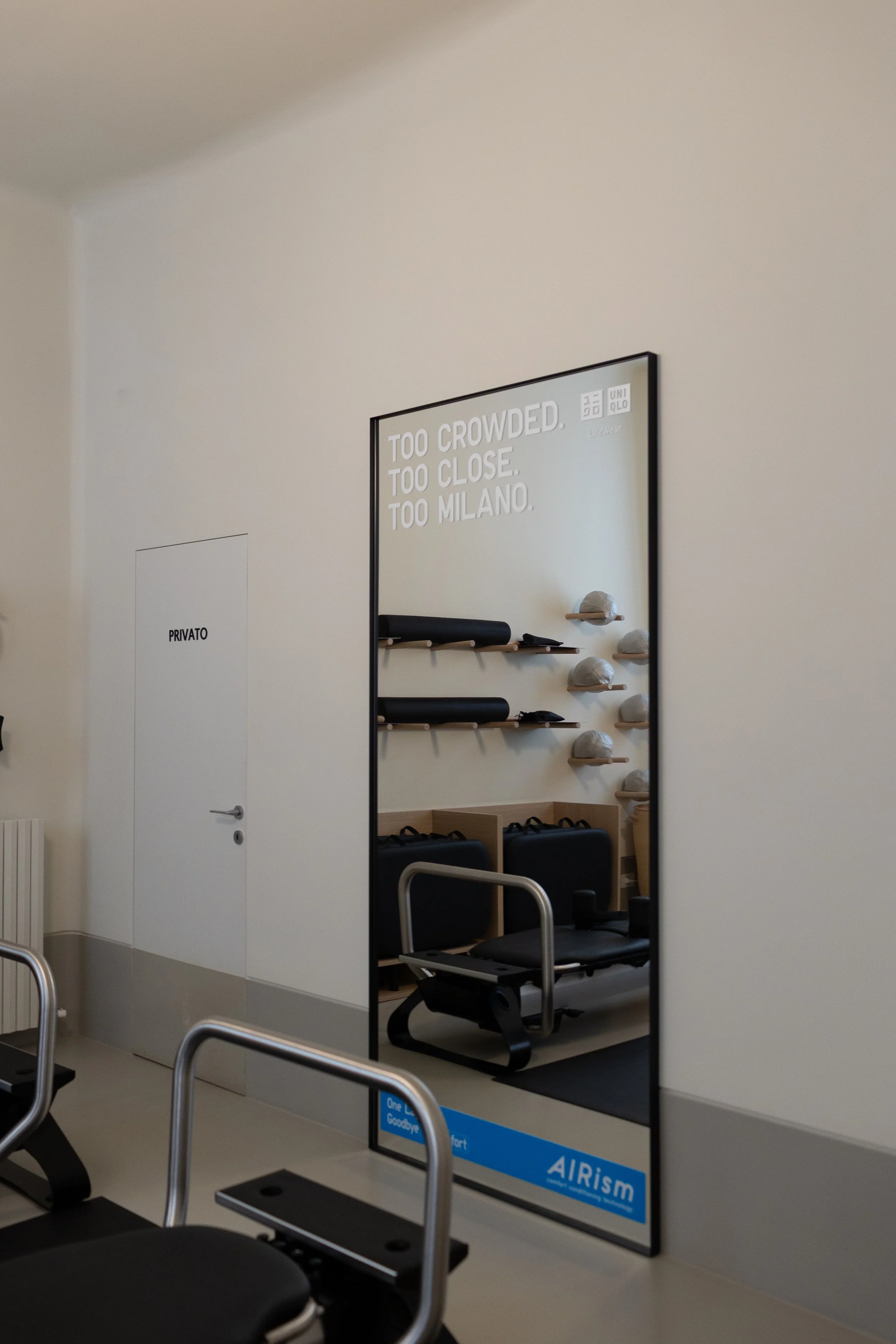 Stanza di reformer pillates con sedie e uno specchio grande con scritta 'Too crowded, too close, too Milano' e scarpe sulla parete. Porta chiusa con scritto 'Privato'.