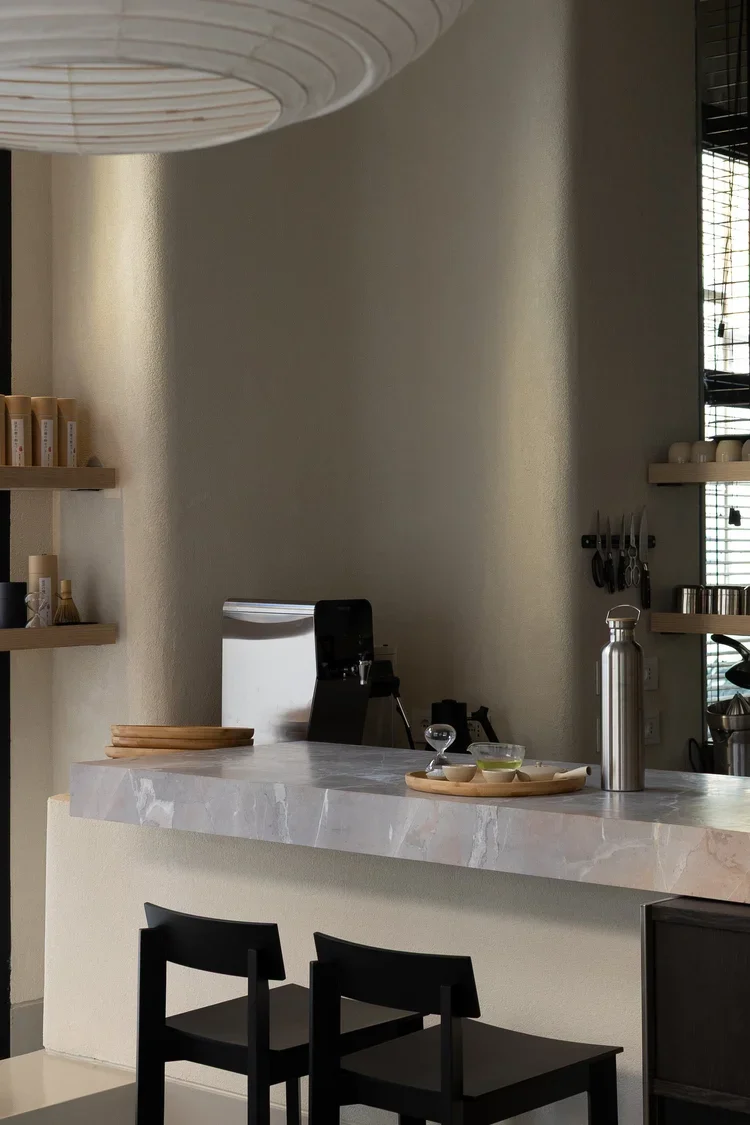 Interno di una locale con bancone in marmo, due sedie nere, oggetti da cucina e scaffali con stoviglie in stile moderno.