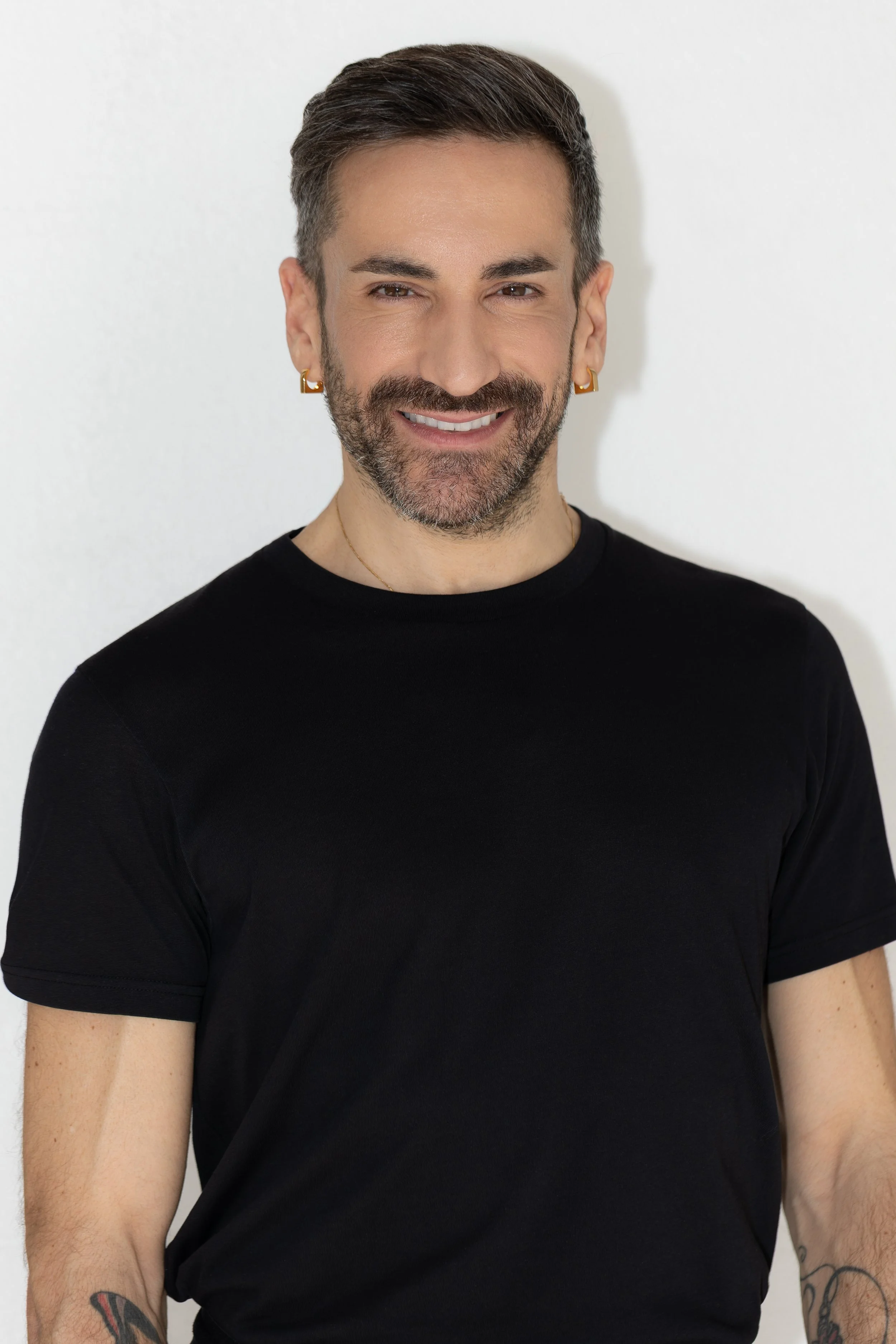 Ritratto di un uomo con barba, occhi castani e capelli castani corti, che indossa orecchini d'oro e una maglietta nera, con sfondo bianco.