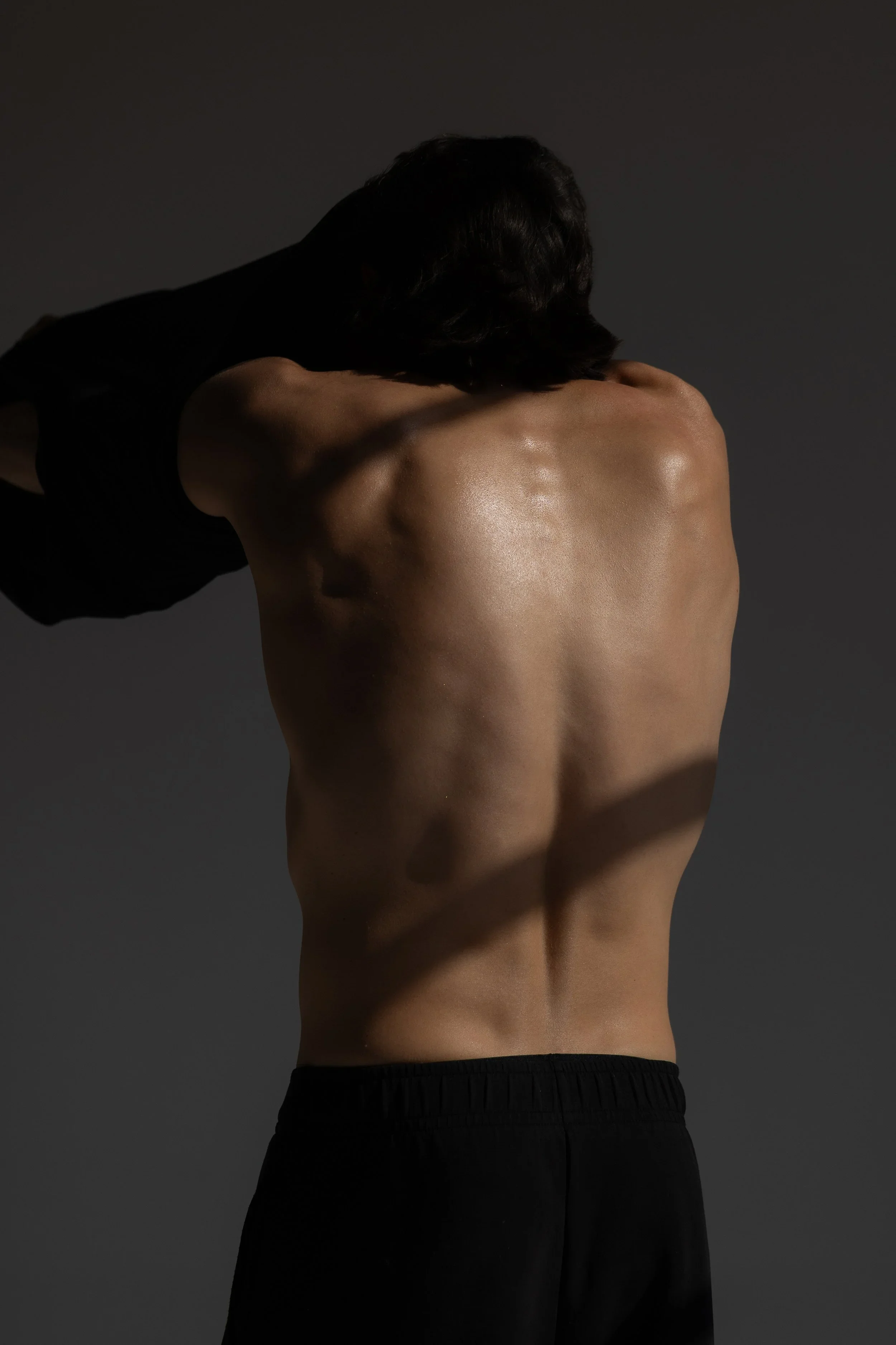 Schiena di una persona con pelle liscia e tonica, con capelli corti e scuri, che indossa pantaloni neri, su sfondo scuro.
