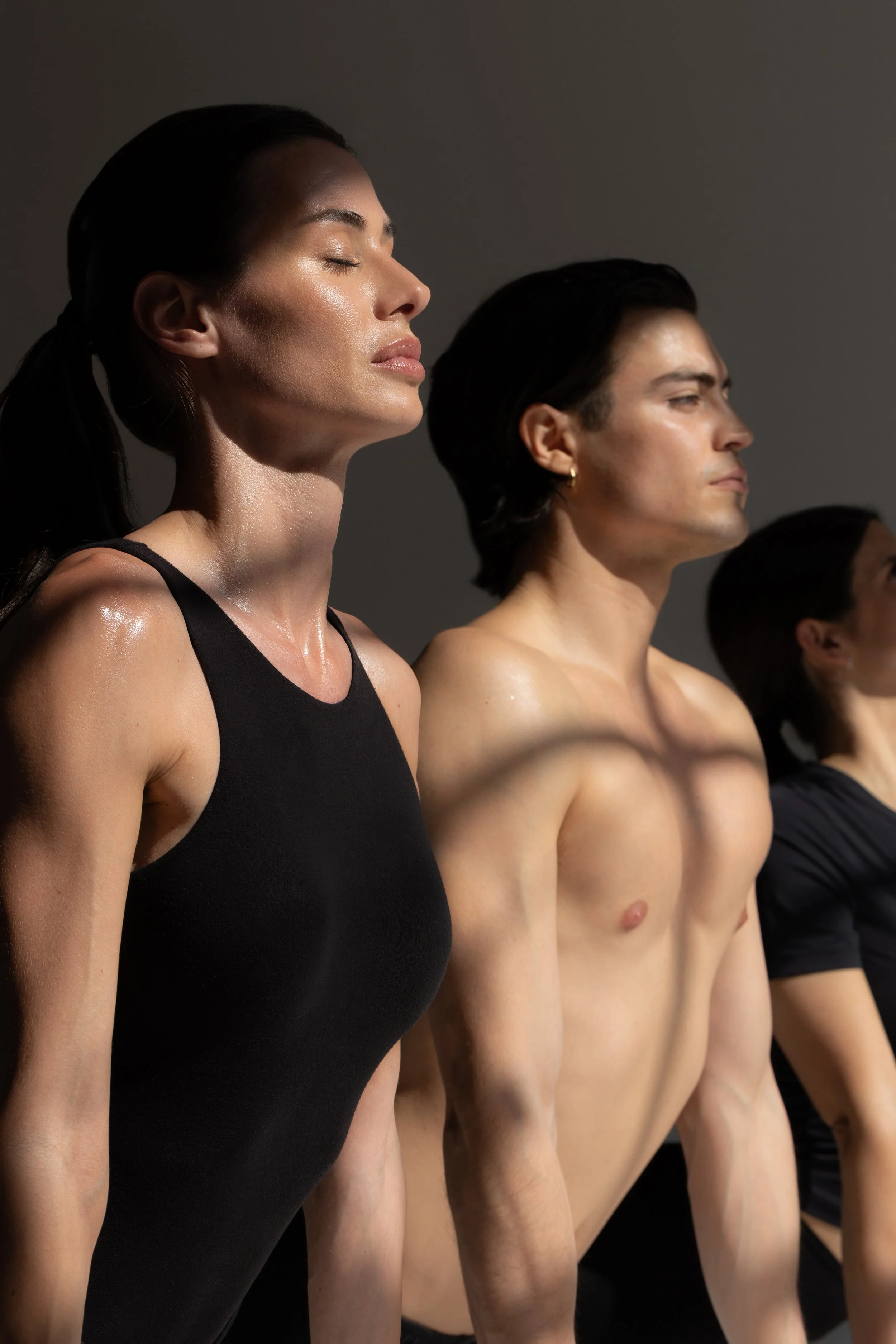 Quattro persone, due donne e due uomini, stanno in piedi in fila, le prime due con gli occhi chiusi, mostrano un atteggiamento di calma e meditazione, indossano abiti scuri, alcune sono senza maglietta, in un ambiente molto semplice con sfondo grigio.