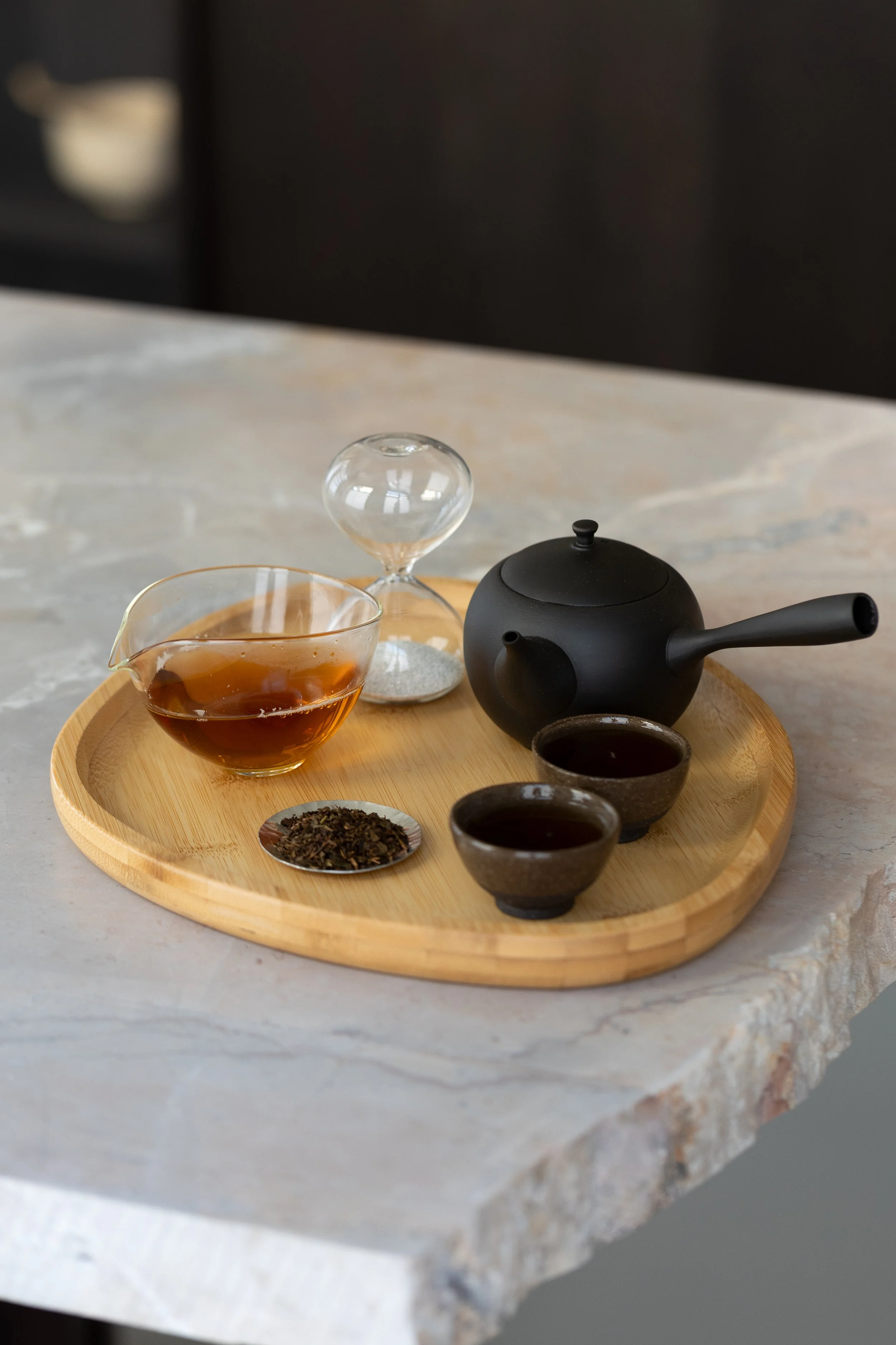 Set di teiere, bicchieri, tazze e tè in diverse forme e colori su un vassoio di bambù, con una superficie di marmo bianco sottostante.