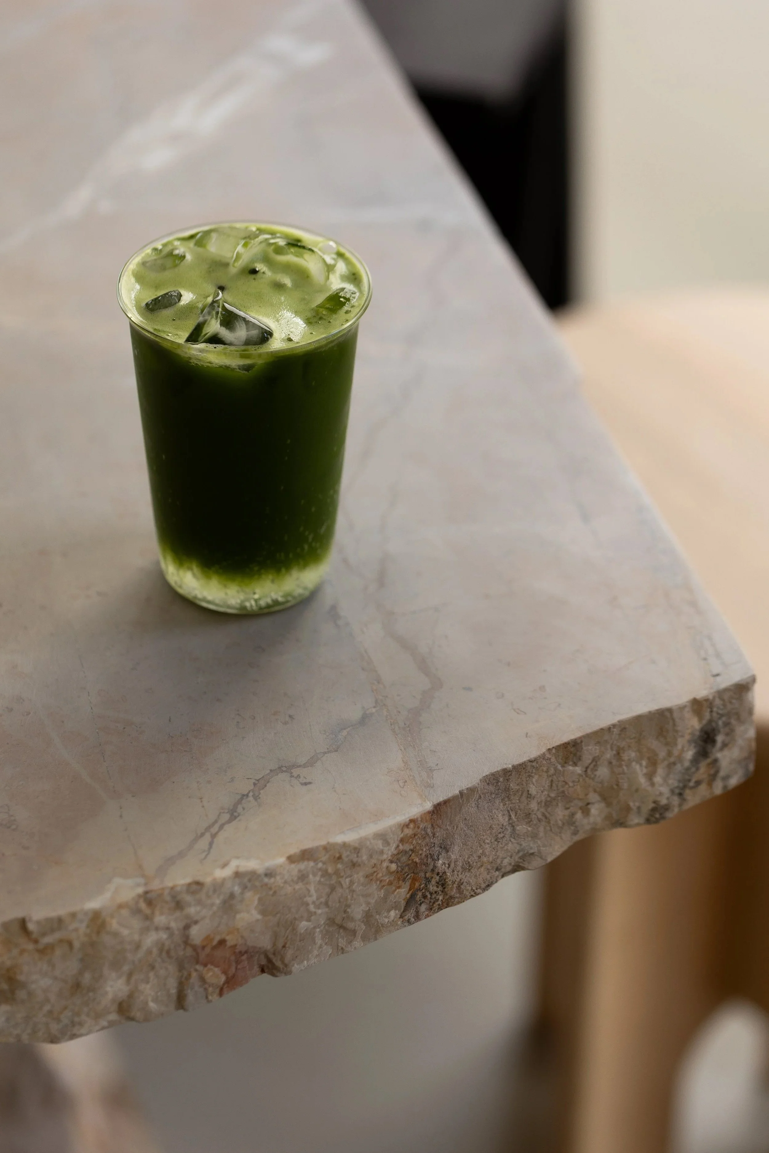 Un bicchiere di vetro trasparente con una bevanda verde, probabilmente matcha, con cubetti di ghiaccio, appoggiato su un tavolo di pietra chiara.