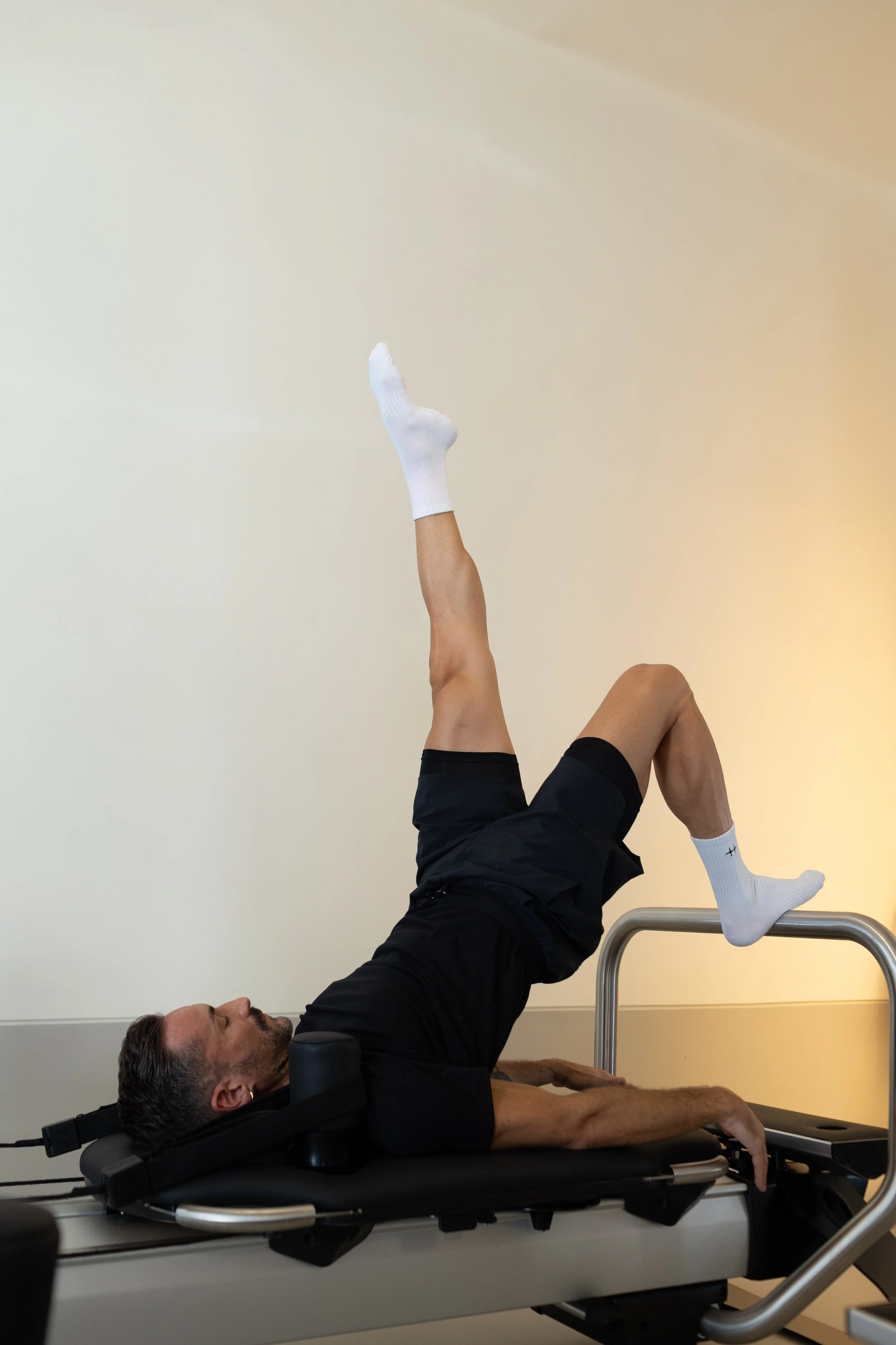 Un uomo disteso su un lettino reformer, con le gambe sollevate e un piede con calzino bianco, mentre tiene un'altra gamba alzata. La scena sembra essere in un ambiente di allenamento.