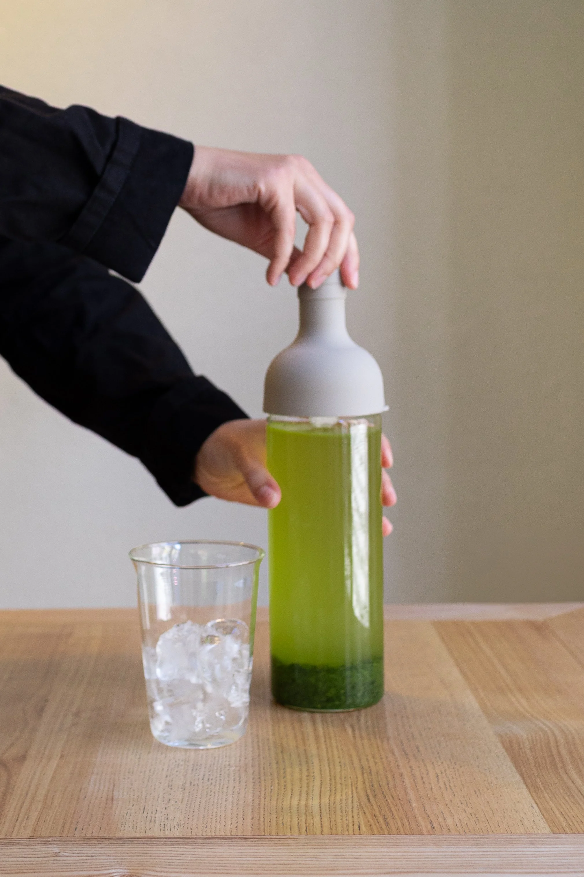Una mano che preme una bottiglia di vetro con liquido verde, bicchiere con acqua e ghiaccio sul tavolo.