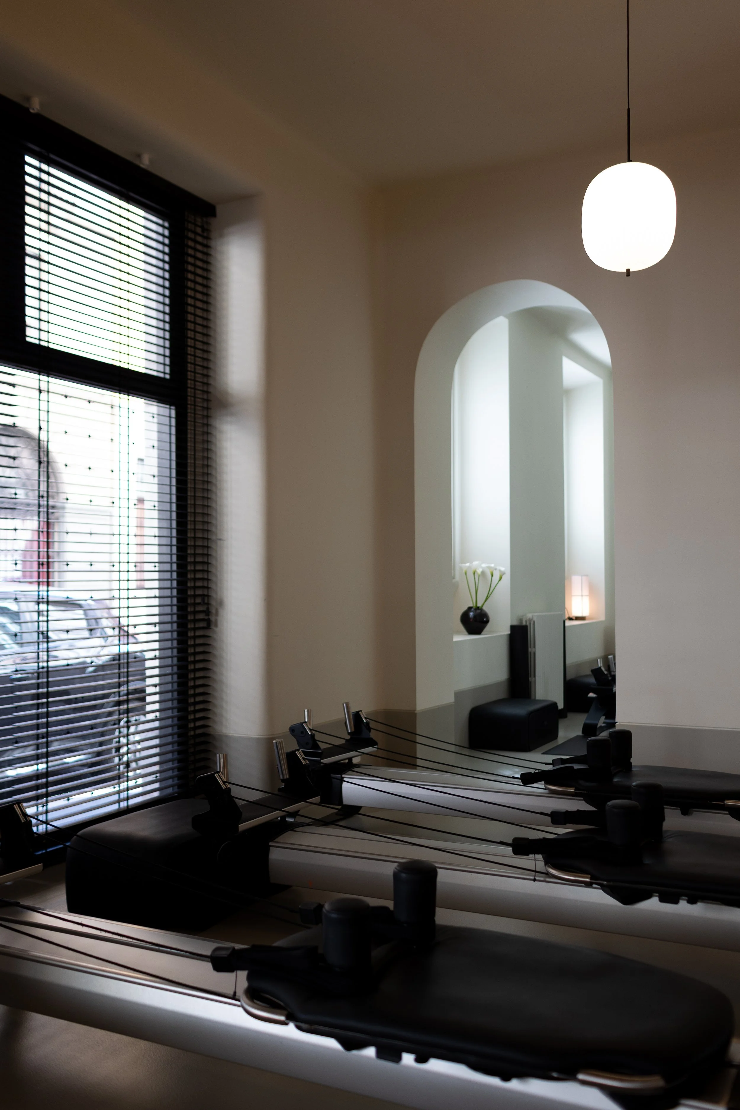 Interno di un barbiere o centro estetico con grandi vetrate con tende a veneziana nere, illuminato da una lampada da soffitto bianca. Sullo sfondo, una nicchia con una pianta e una lampada, e alcuni strumenti per parrucchieri o estetisti sono posizionati sui banconi.