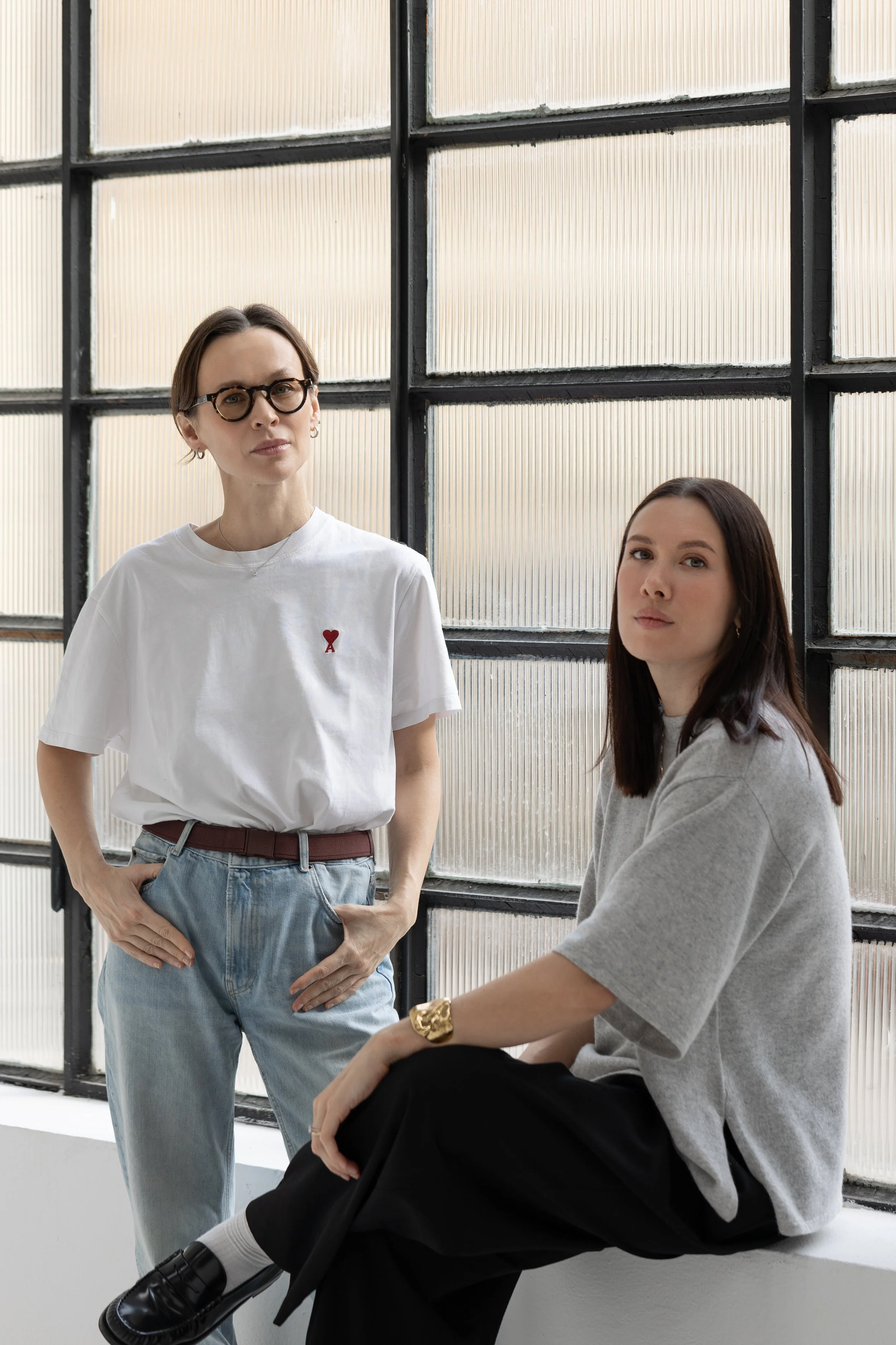 Due donne in un ambiente luminoso davanti a una grande finestra con vetri oscurati, una in piedi con occhiali tondi, maglietta bianca con cuore rosso, e pantaloni larghi, l'altra seduta con maglione grigio, pantaloni scuri e accessori dorati.