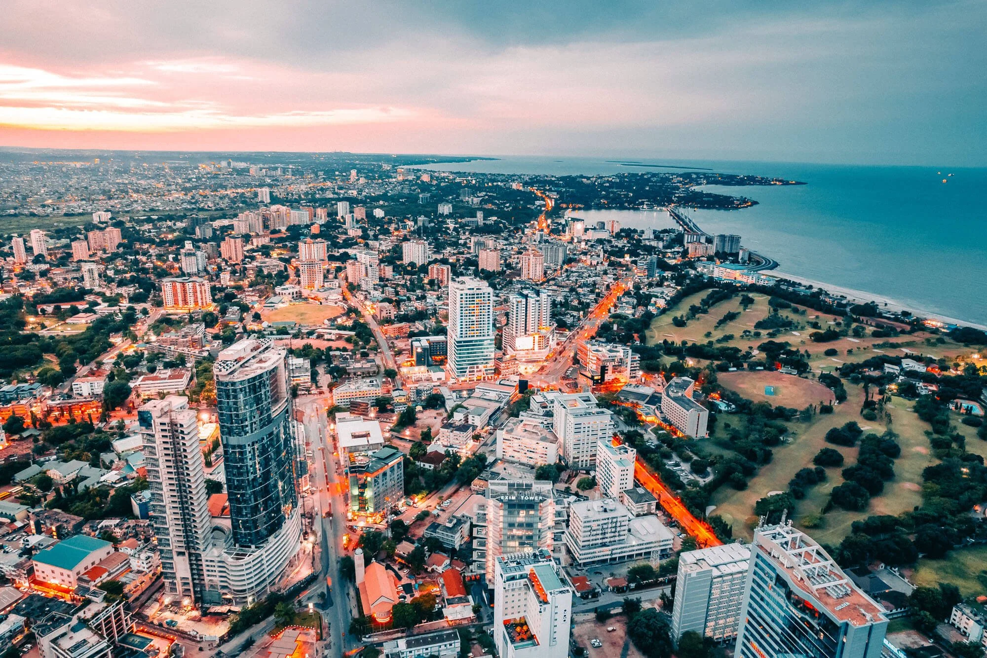 An aerial view of a cityscape during sunset, featuring modern high-rise buildings, a park, and a coastline in the distance.