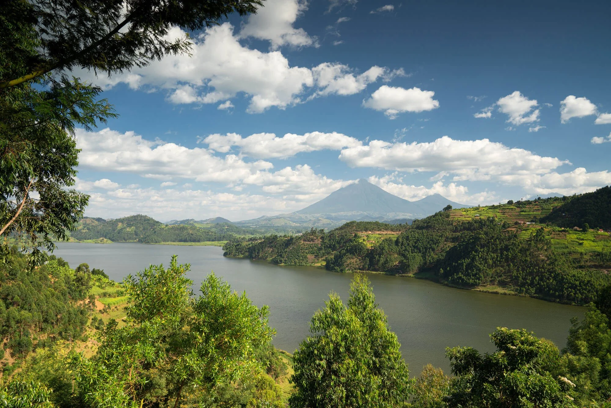 A scenic landscape with a river flowing through lush green hills, with a mountain in the background under a blue sky with scattered clouds.