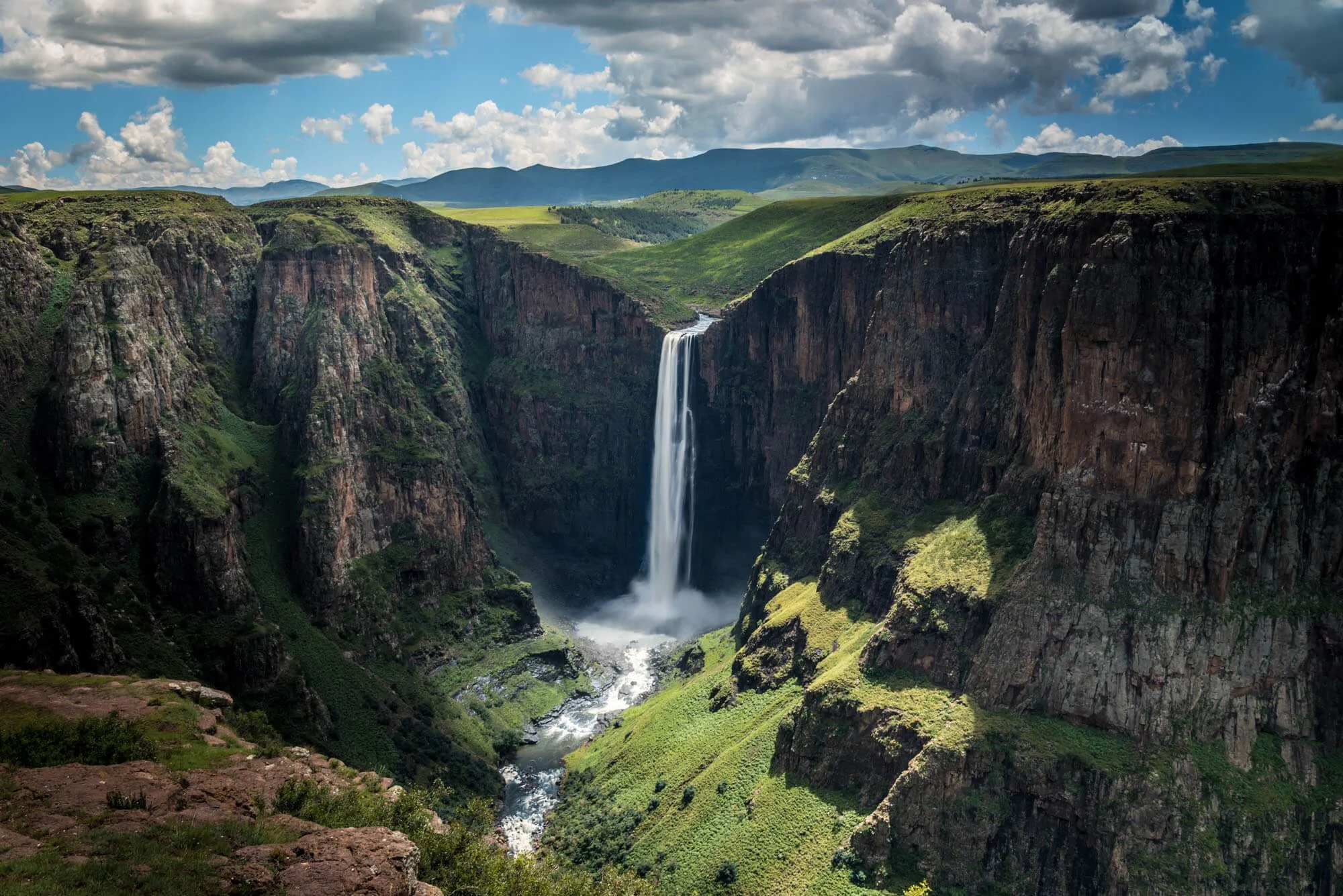 A tall waterfall cascading down a deep canyon surrounded by green cliffs and hills under a partly cloudy sky.