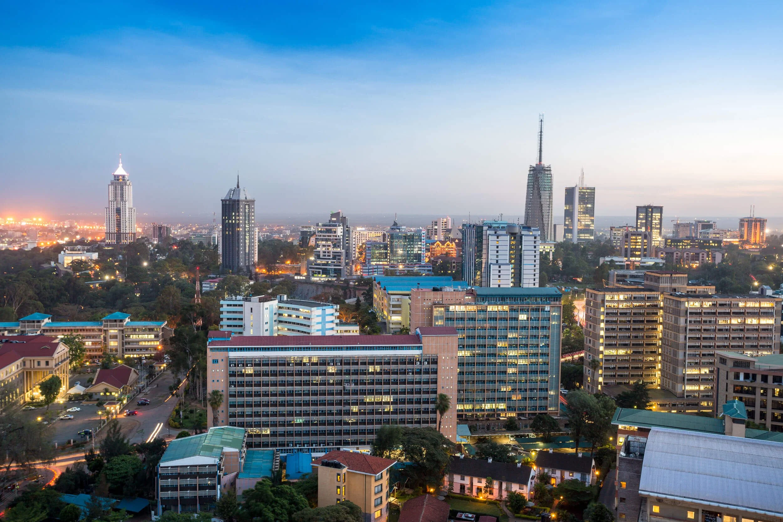 A city skyline at dusk with tall buildings, some lit up, and a mix of modern and older architecture, with the sky transitioning from blue to light.