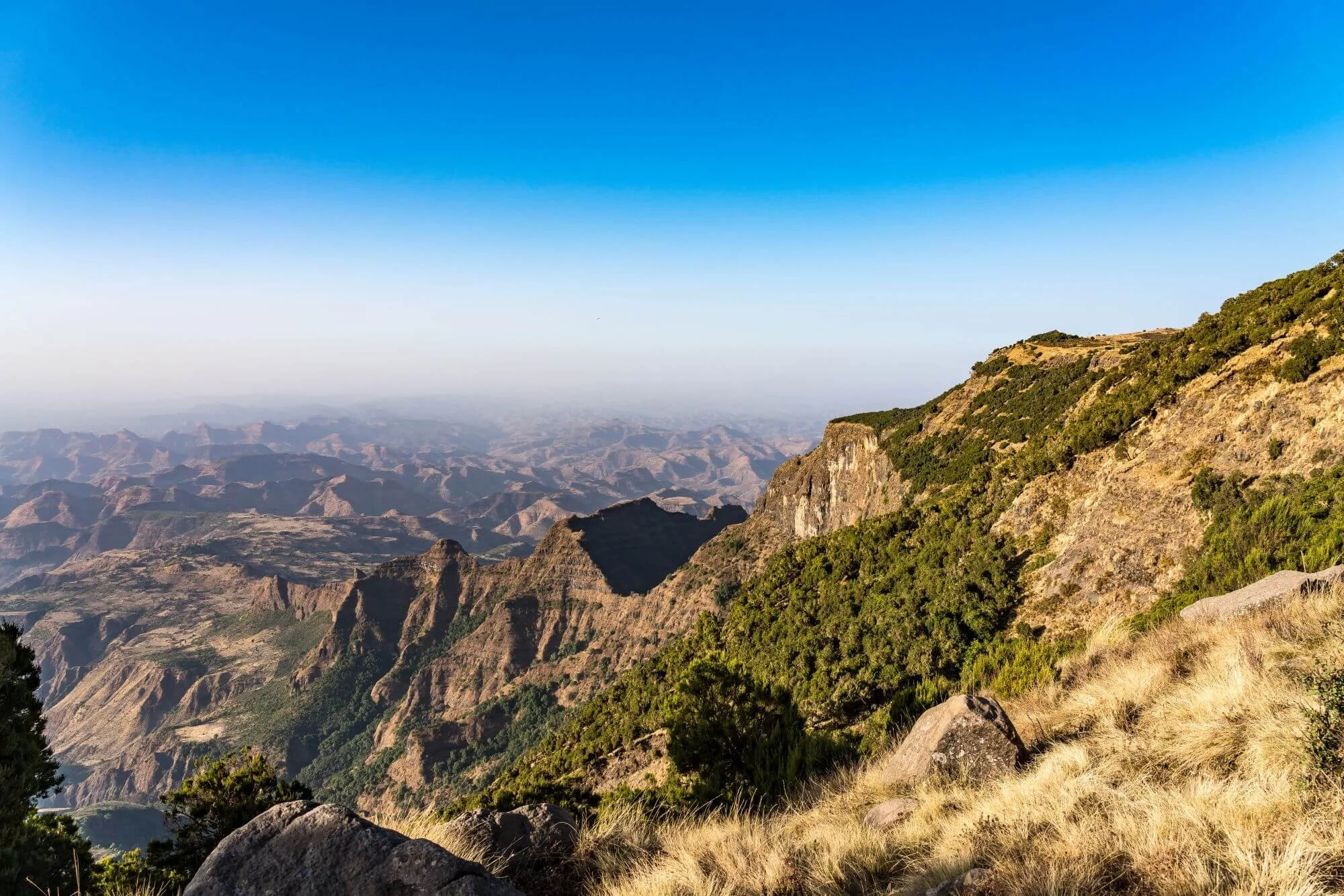Mountain landscape with rugged cliffs, green shrubs on the slopes, and a clear blue sky overhead.