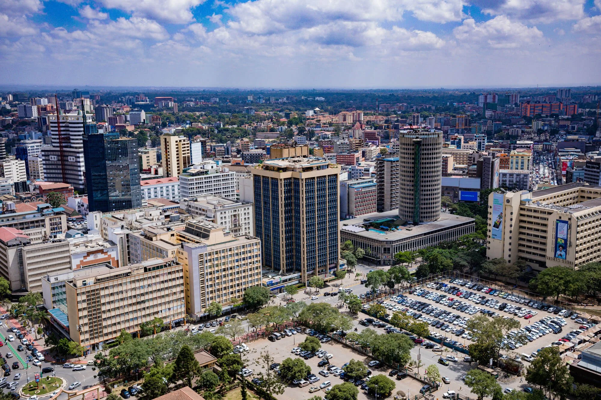 Aerial view of a city skyline with tall buildings, roads, and scattered trees under a partly cloudy sky.