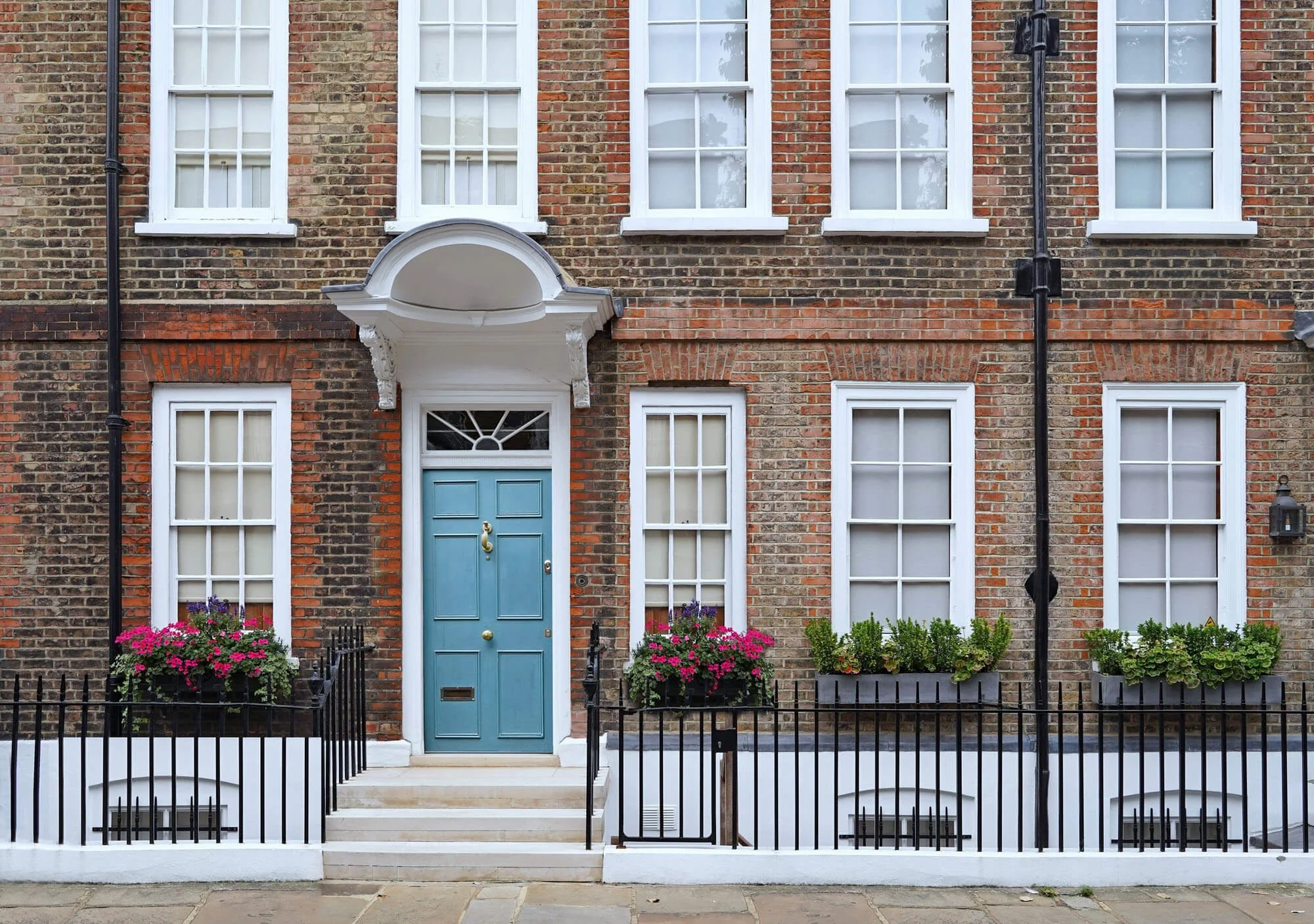 A brick townhouse with white window trim, a blue front door, flower boxes with pink and purple flowers, and black iron fencing.
