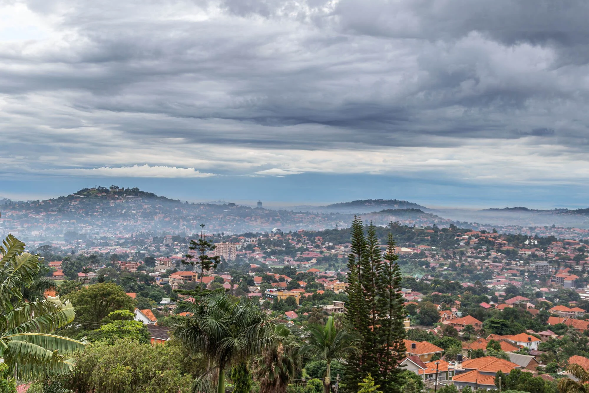 A cityscape with residential houses and trees in the foreground, hills in the background, and a cloudy sky overhead.