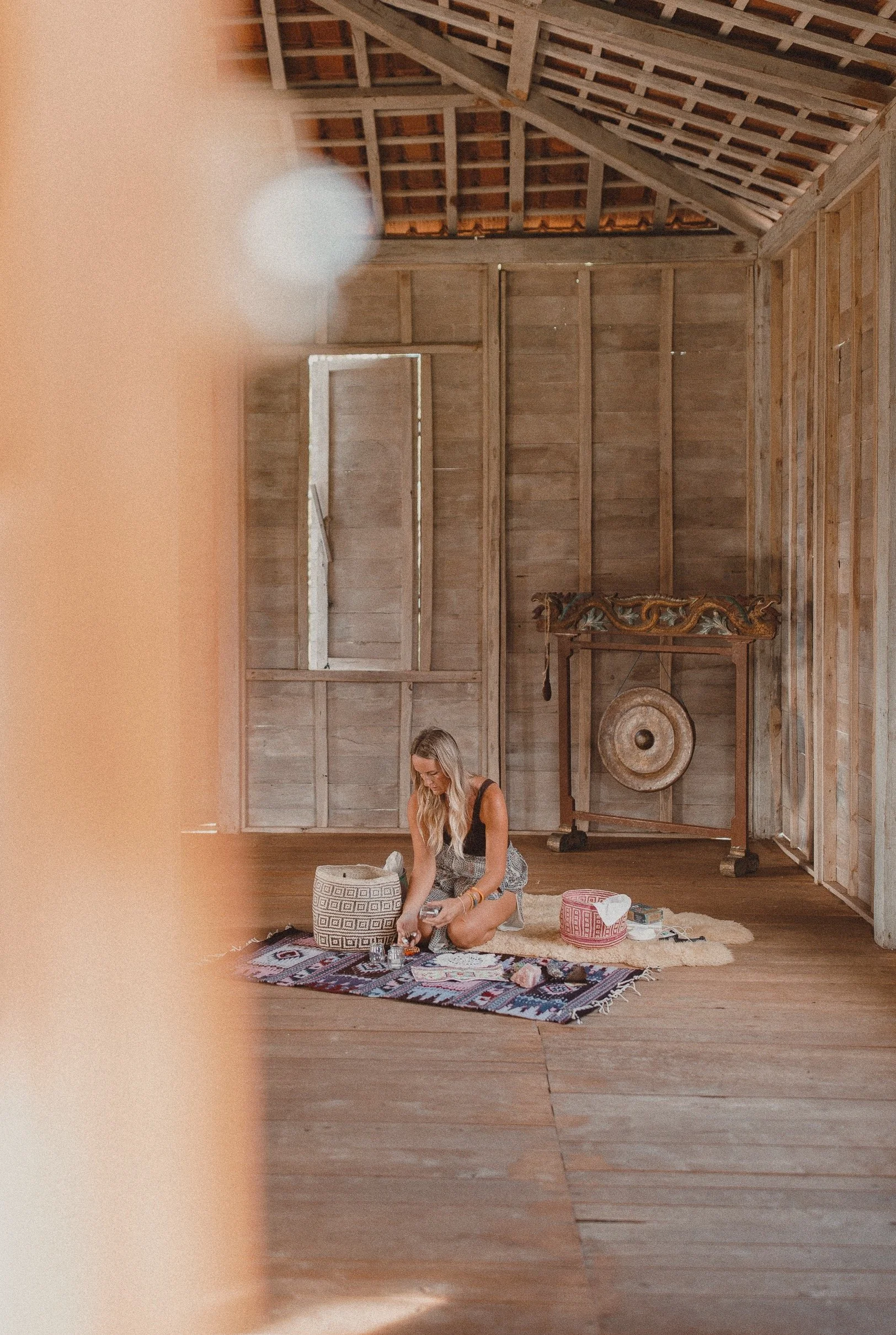 A woman is sitting on a colorful rug inside a wooden room with unfinished walls and ceiling. She is organizing items in a basket, with a gong and a drum in the background.