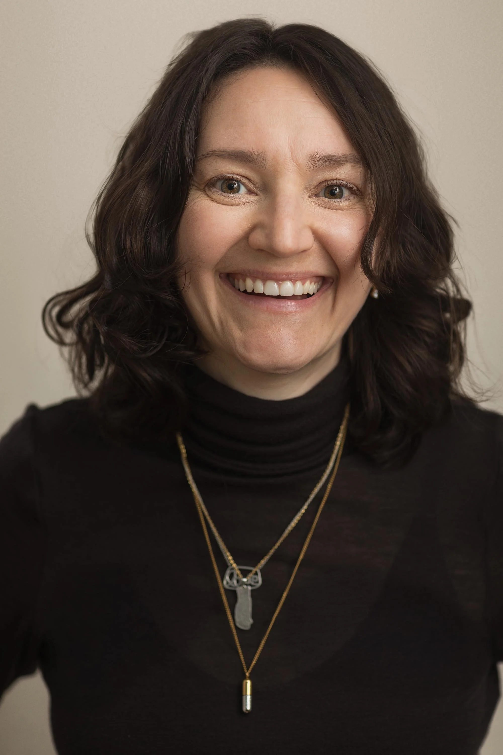A woman with shoulder-length dark hair, wearing a black turtleneck and gold necklaces, smiling at the camera against a plain background.
