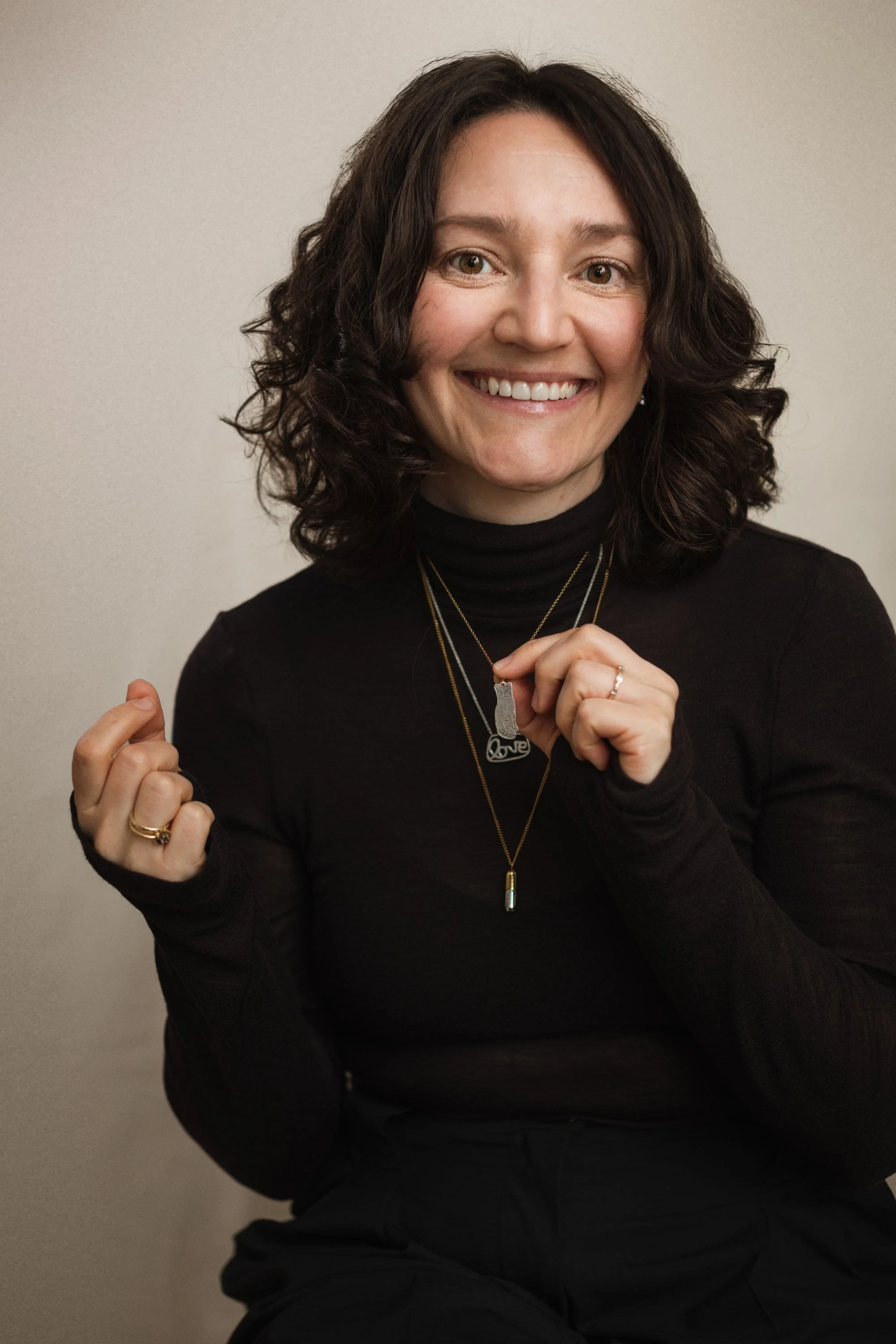 A woman with dark wavy hair smiling and holding up multiple necklaces, dressed in black and against a neutral background.