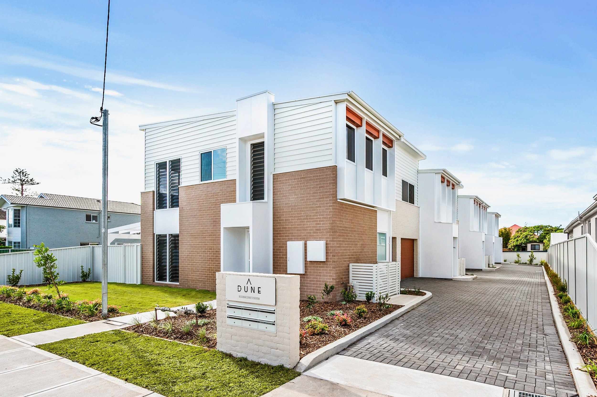 Modern residential building with white and brick exterior, landscaped front yard, and a sign that reads 'Dune' in front of the property. Clear blue sky above.