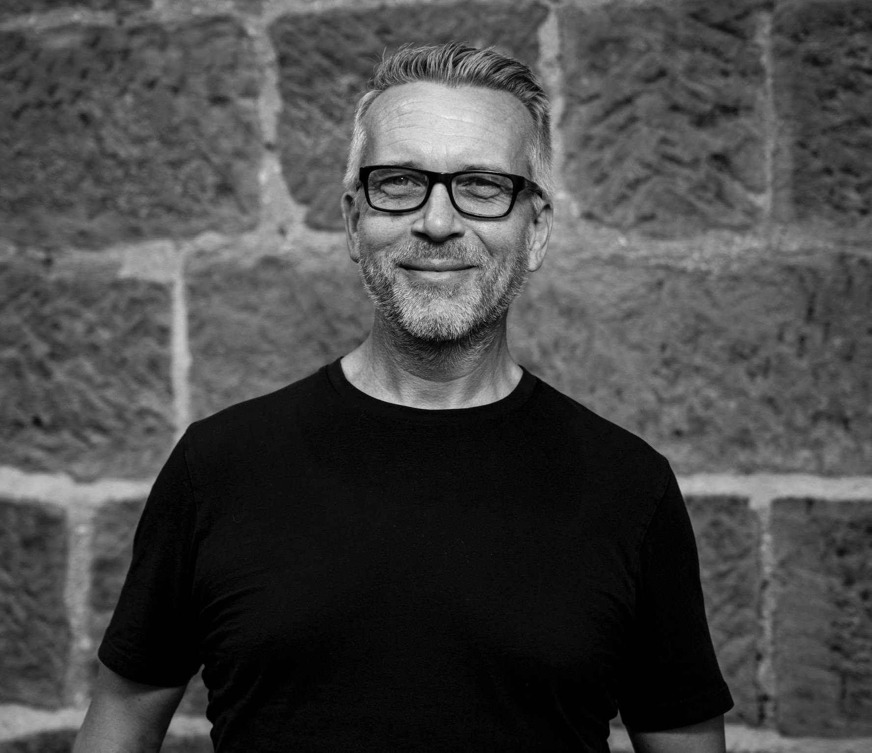 A smiling middle-aged man with glasses and a beard standing in front of a brick wall, wearing a black shirt.