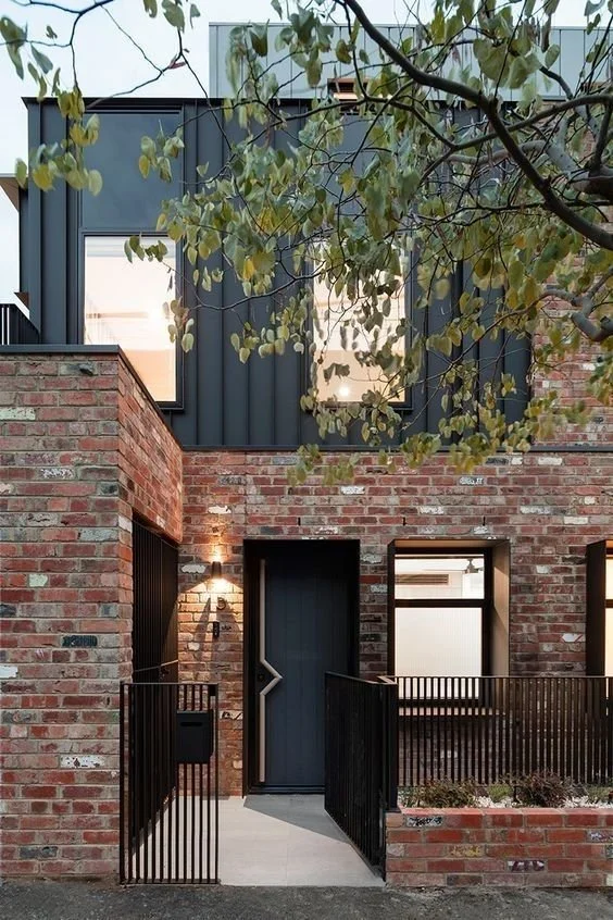 Modern brick house with black metal accents and large windows, surrounded by green tree leaves.
