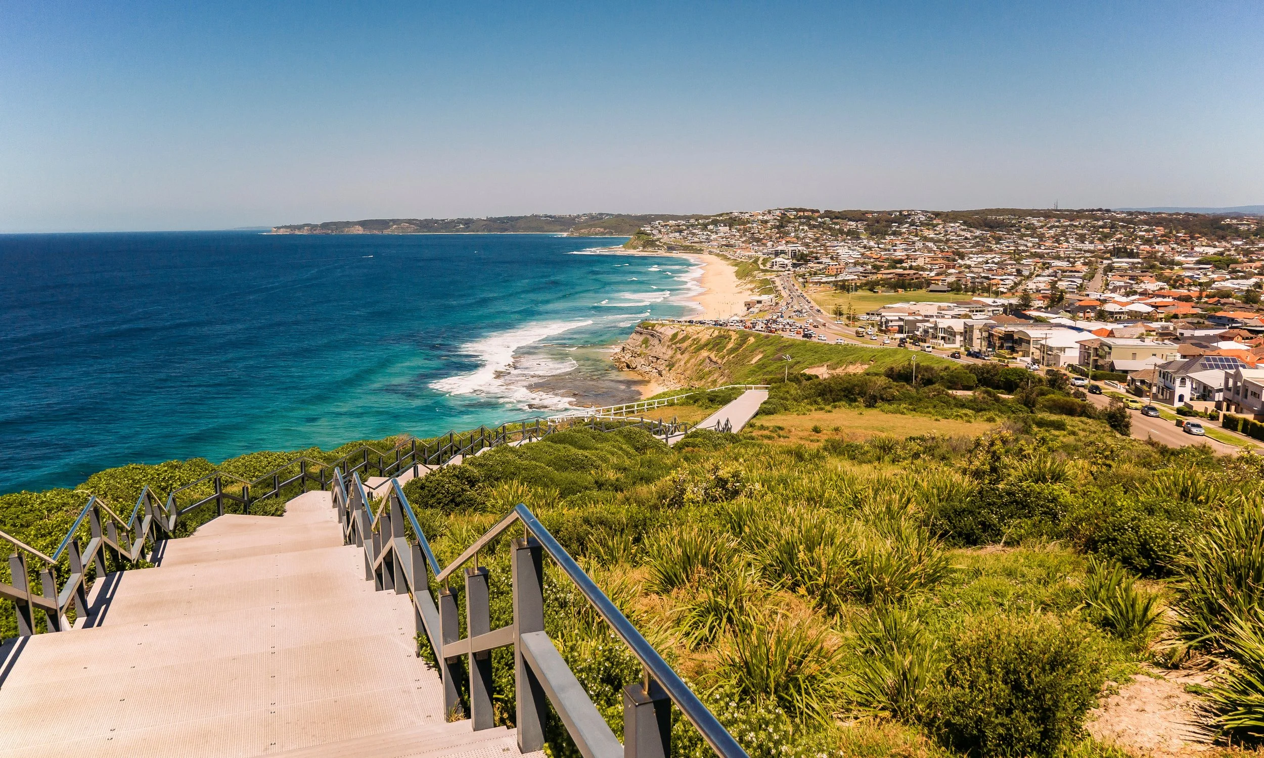 Coastal view with stairs leading down to a beach, surrounded by green vegetation, and a town with houses and buildings on the hillside under a clear blue sky.