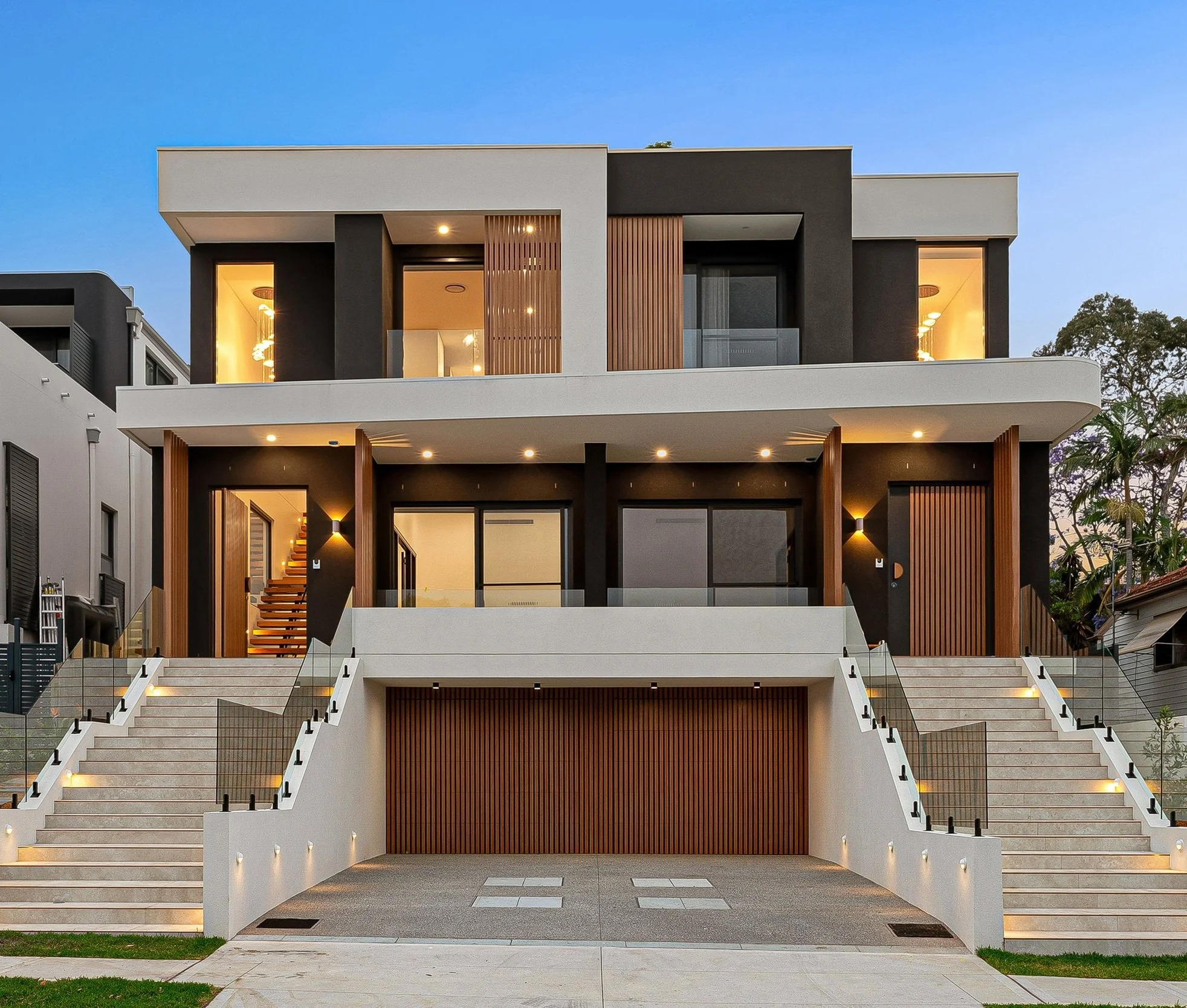 Modern multi-story house with white and black exterior walls, large windows with warm interior lighting, and wooden accents. Front staircase with multiple steps leads to entrance, with outdoor lighting along stairs and ground level. Trees and neighboring buildings are visible.