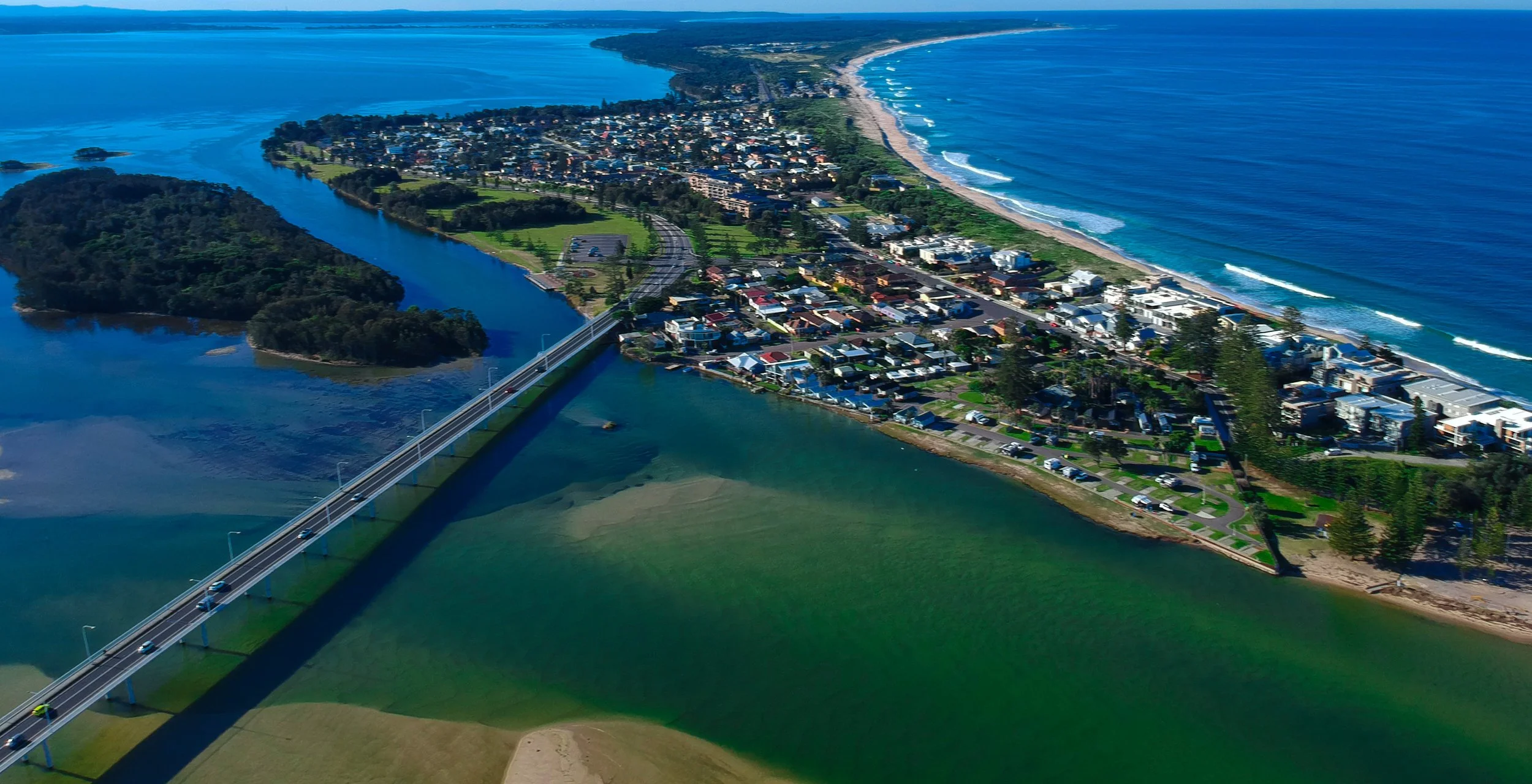 An aerial view of a coastal town with a bridge over a river, a green park, residential houses, and a beach along the ocean with waves.