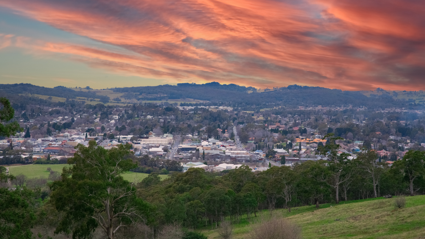 A panoramic view of a town at sunset with colorful clouds in the sky, green rolling hills in the foreground, and buildings and trees spread across the town.